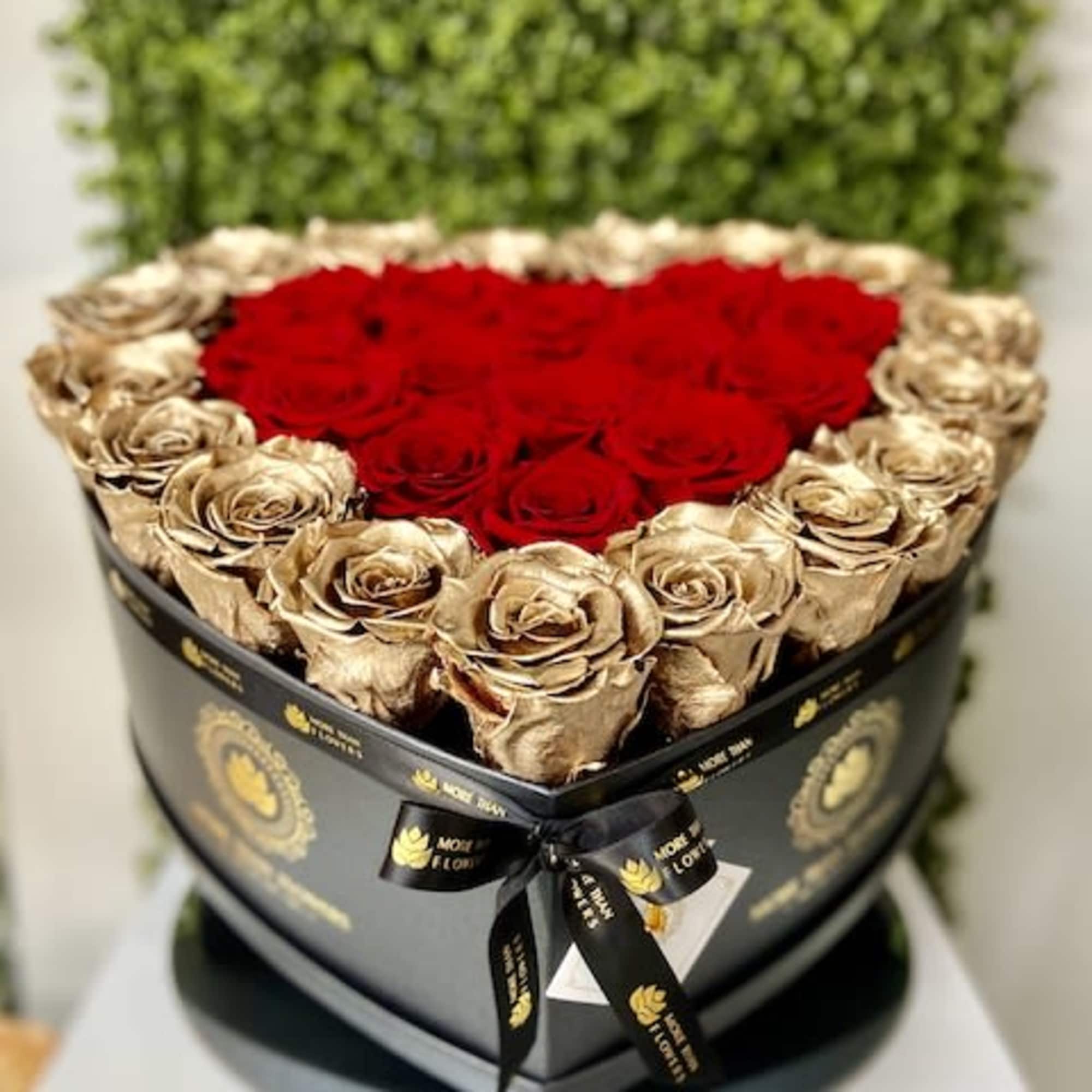 Heart-shaped black box of red and gold roses with logo ribbon against a green backdrop