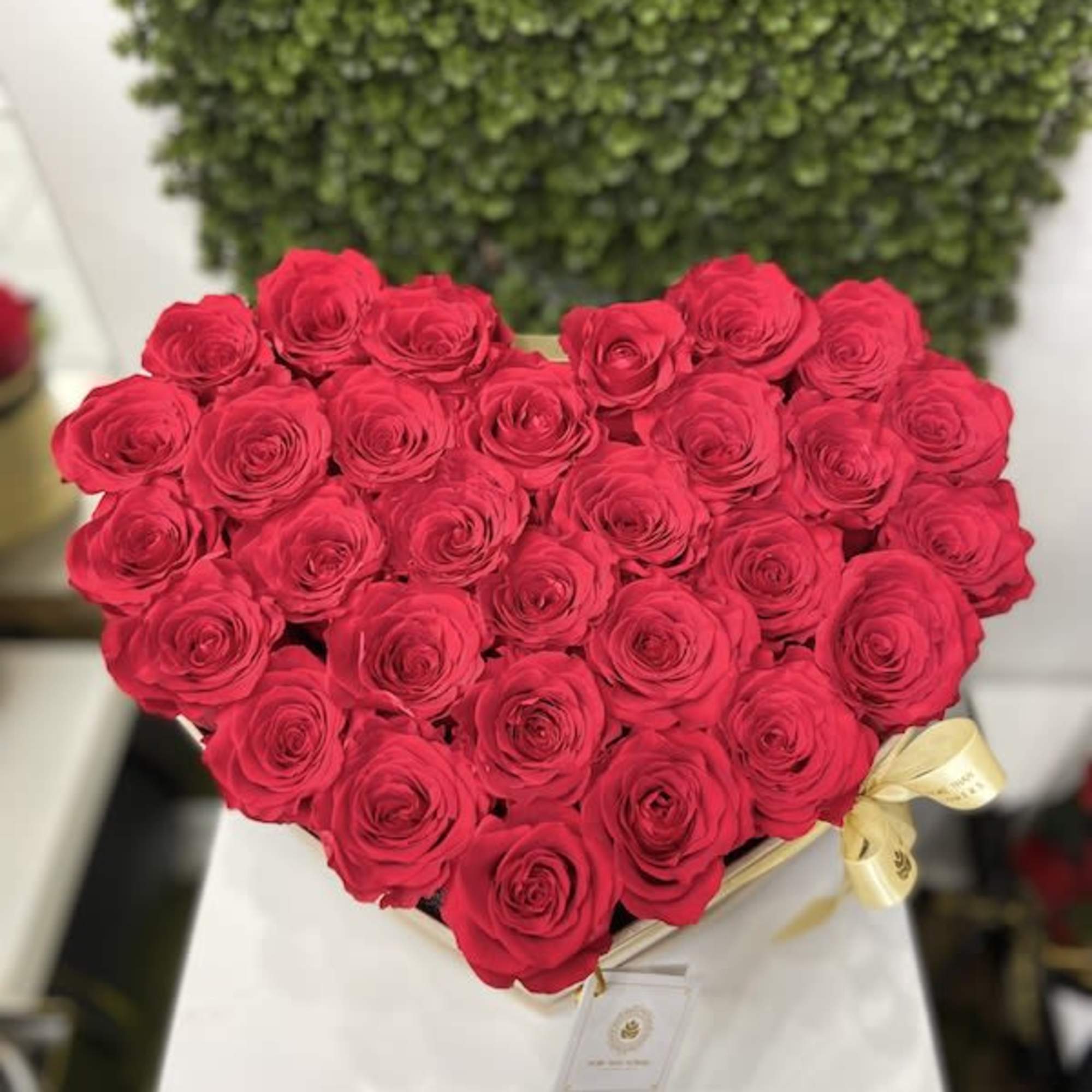Heart-shaped arrangement of red roses in a box with a gold ribbon on a white pedestal.