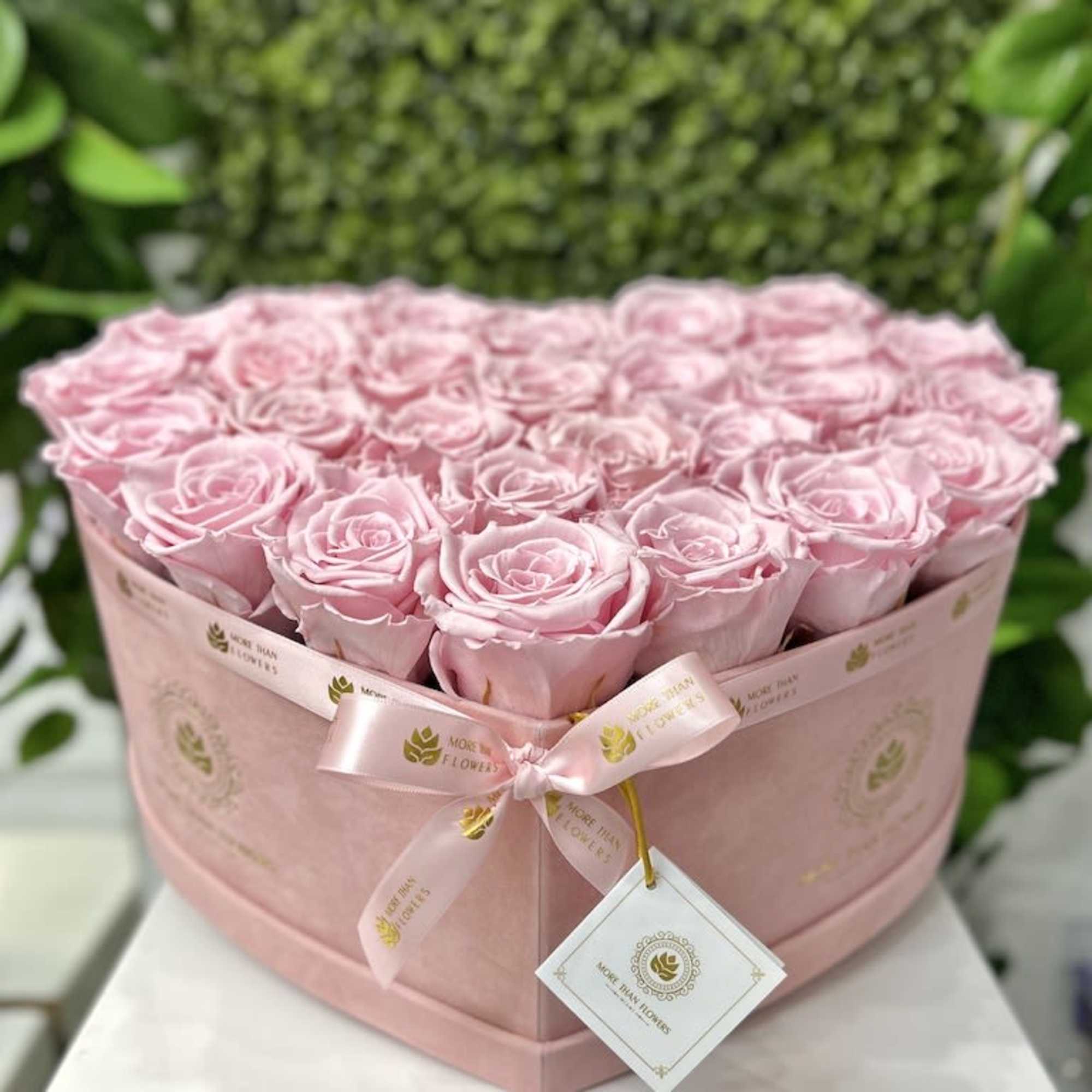 Heart-shaped pink box filled with tightly packed light pink roses and a satin ribbon tag.