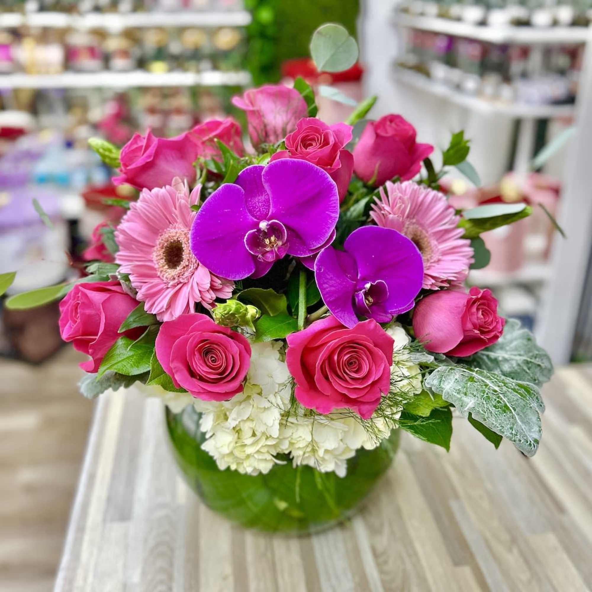 Low round bouquet of hot pink roses, pink gerbera daisies, purple orchids, and white hydrangeas in a glass bowl vase