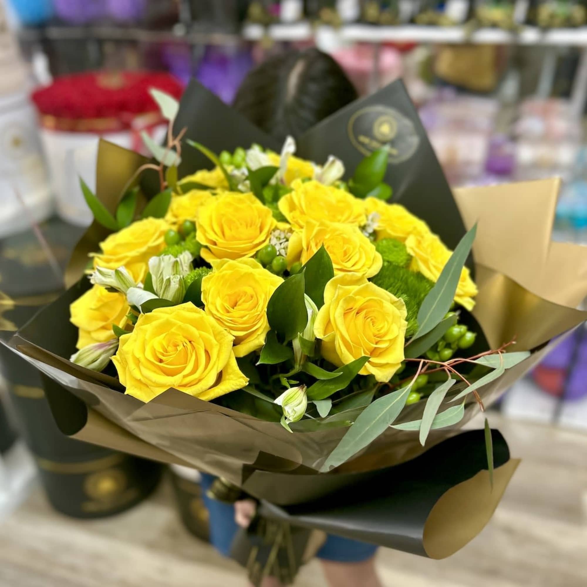 Hand-tied bouquet of bright yellow roses with white blooms and greenery in black and gold wrap
