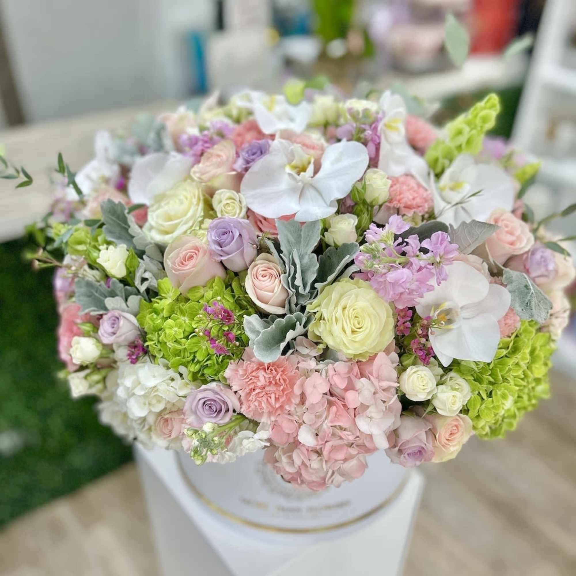 Round pastel arrangement of roses, hydrangeas, orchids and carnations in a white hatbox.
