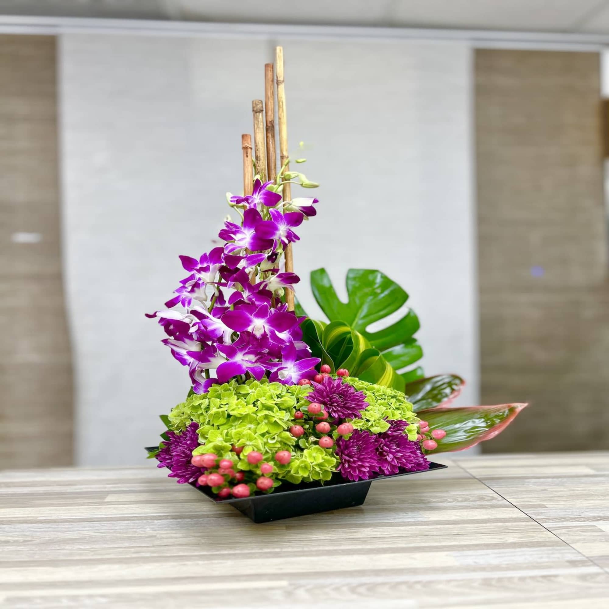 Modern arrangement with purple orchids, green hydrangeas, and mums in a low black container with tall bamboo.