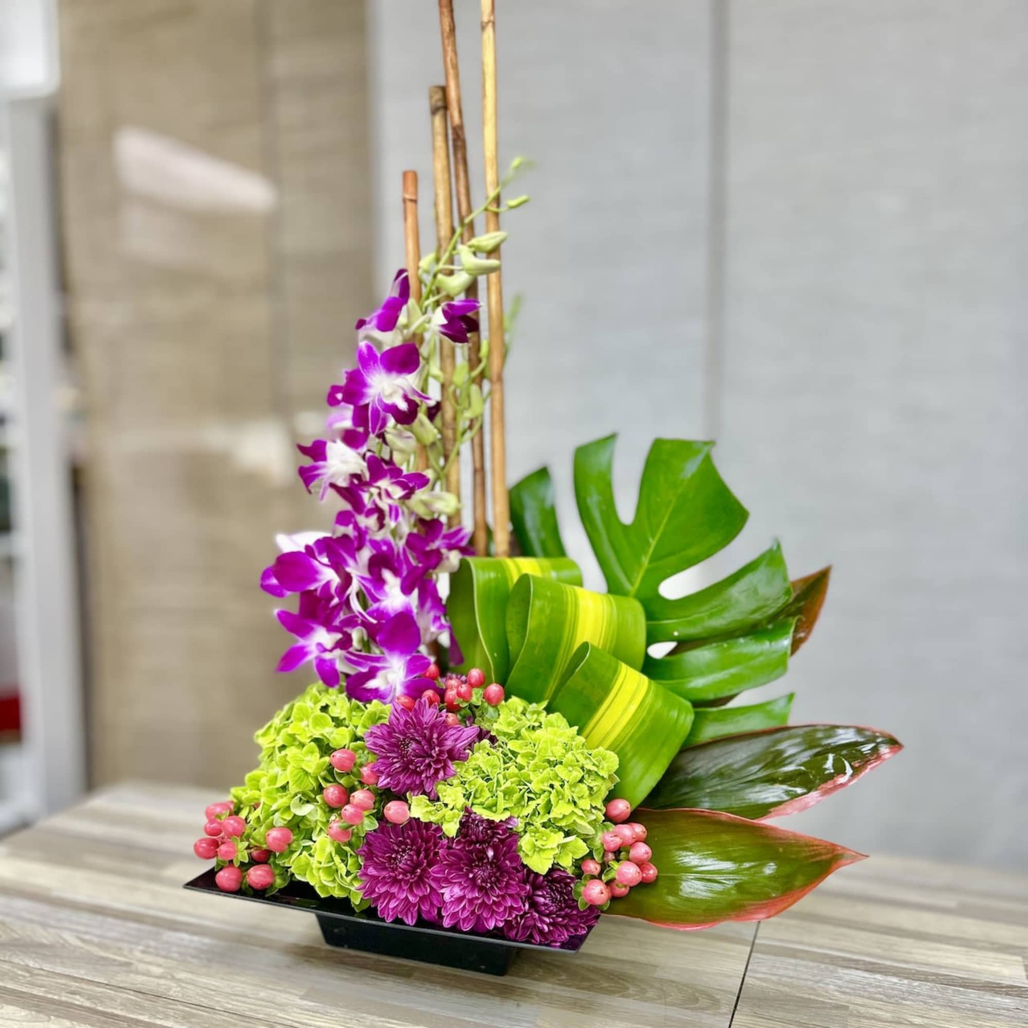 Modern arrangement with purple orchids, green hydrangeas, mums and large tropical leaves in a low black container