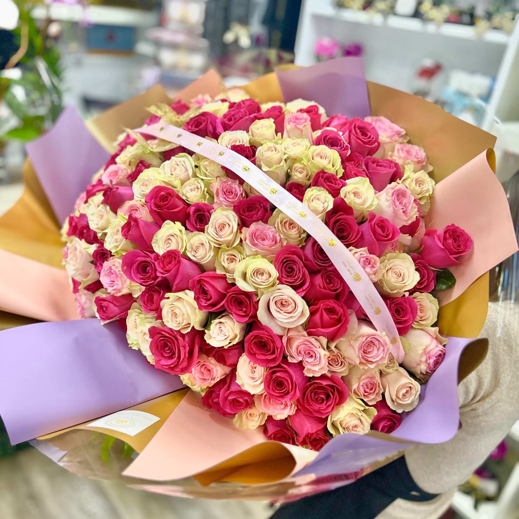 Large round bouquet of hot pink, light pink, and cream roses wrapped in multicolored paper.