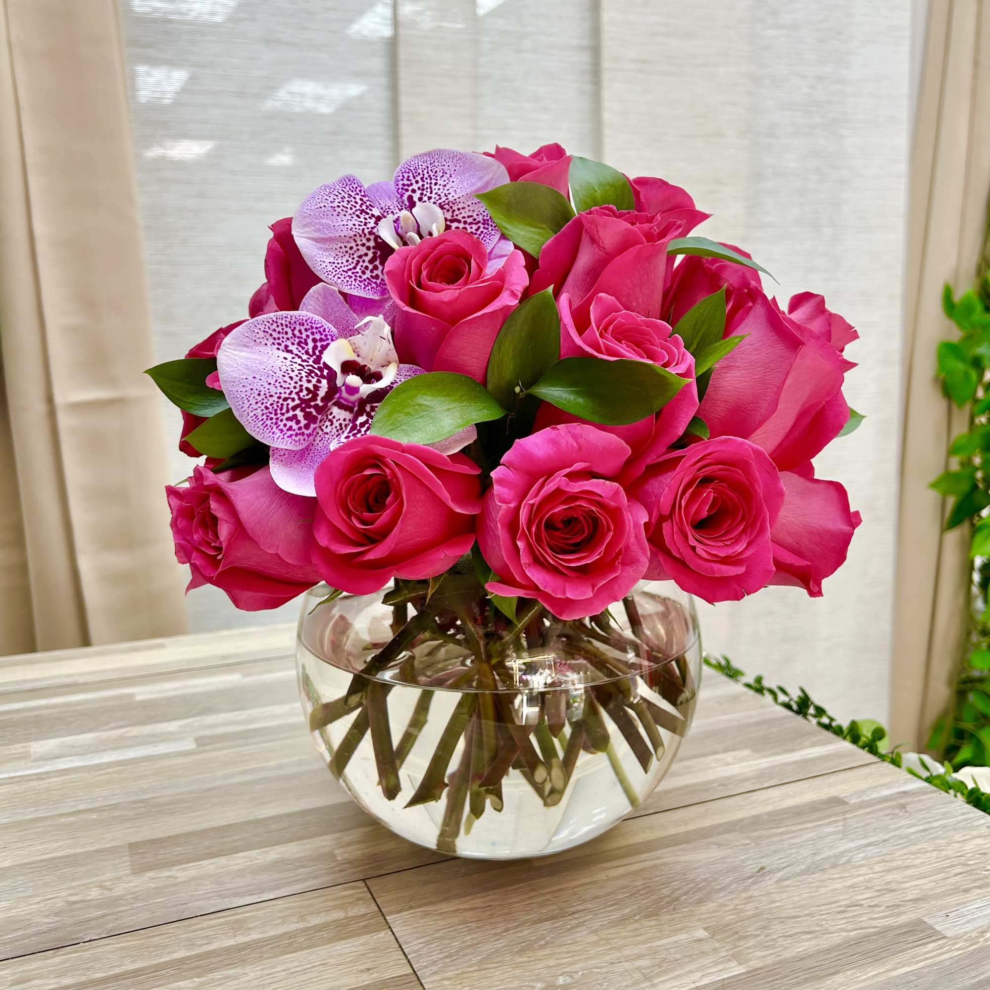 Round arrangement of hot pink roses with speckled purple orchids in a clear glass bowl vase
