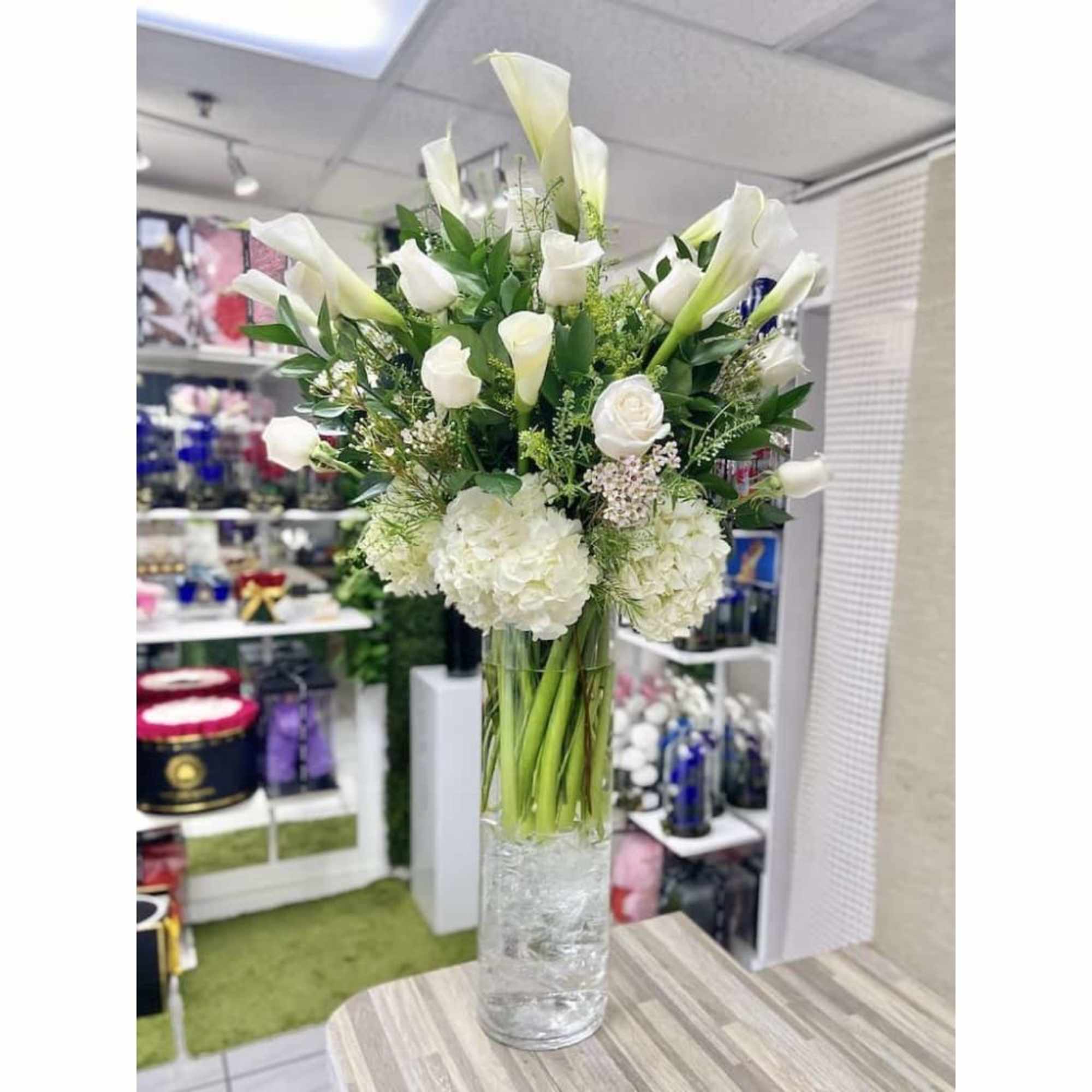 Tall arrangement of white calla lilies, roses, and hydrangeas in a clear glass cylinder vase