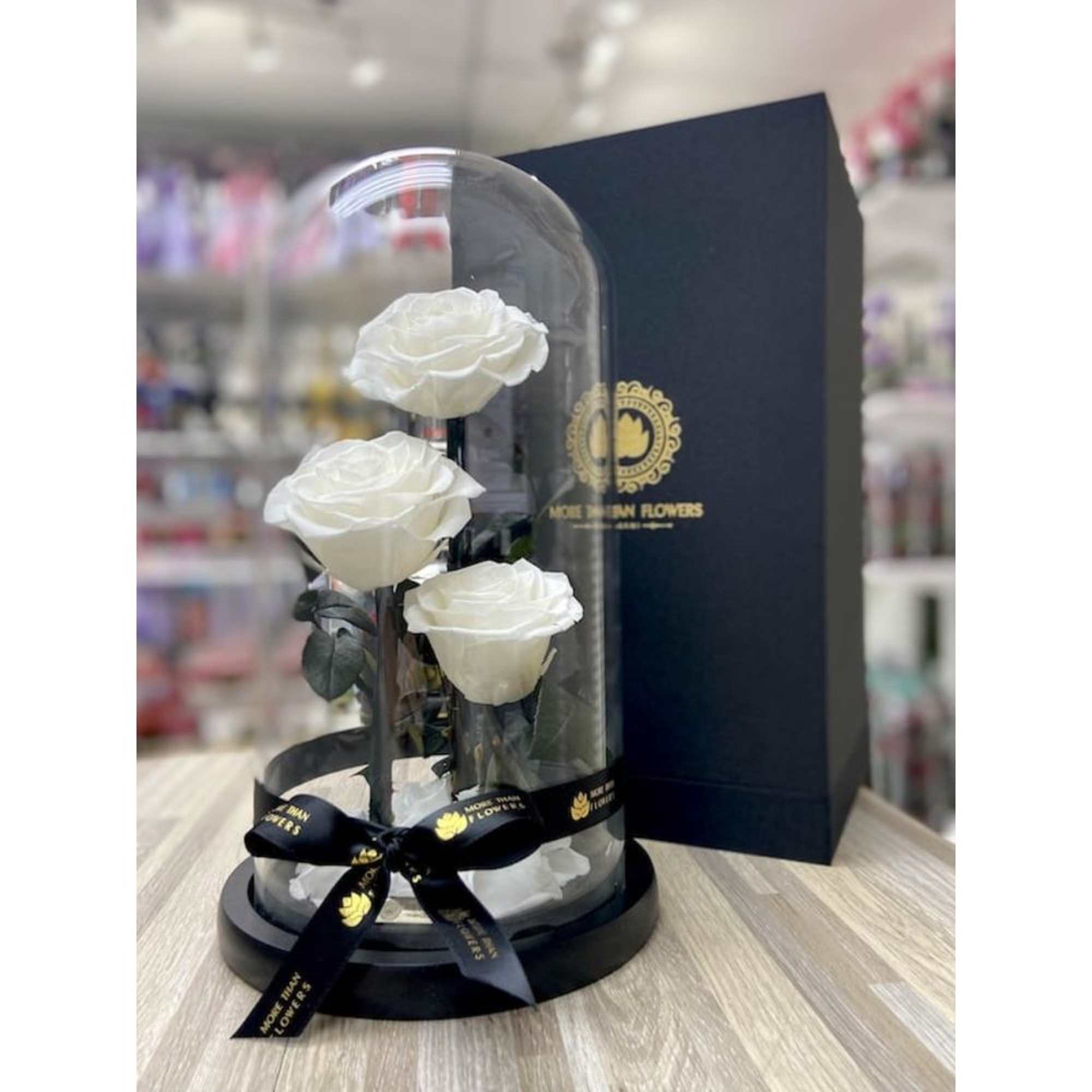 Three white roses displayed under a clear glass dome on a black base with a gift box behind.