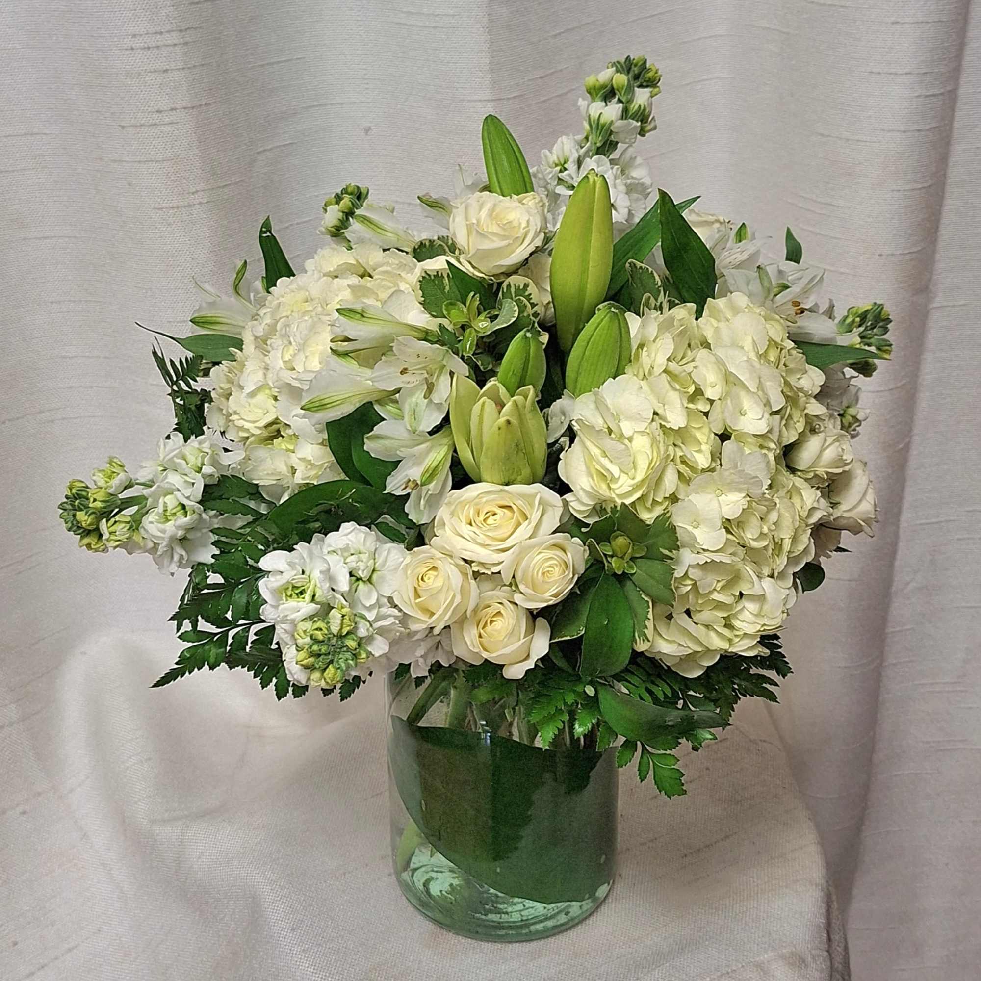 White roses, lilies, hydrangeas, and stock arranged in a clear glass cylinder vase