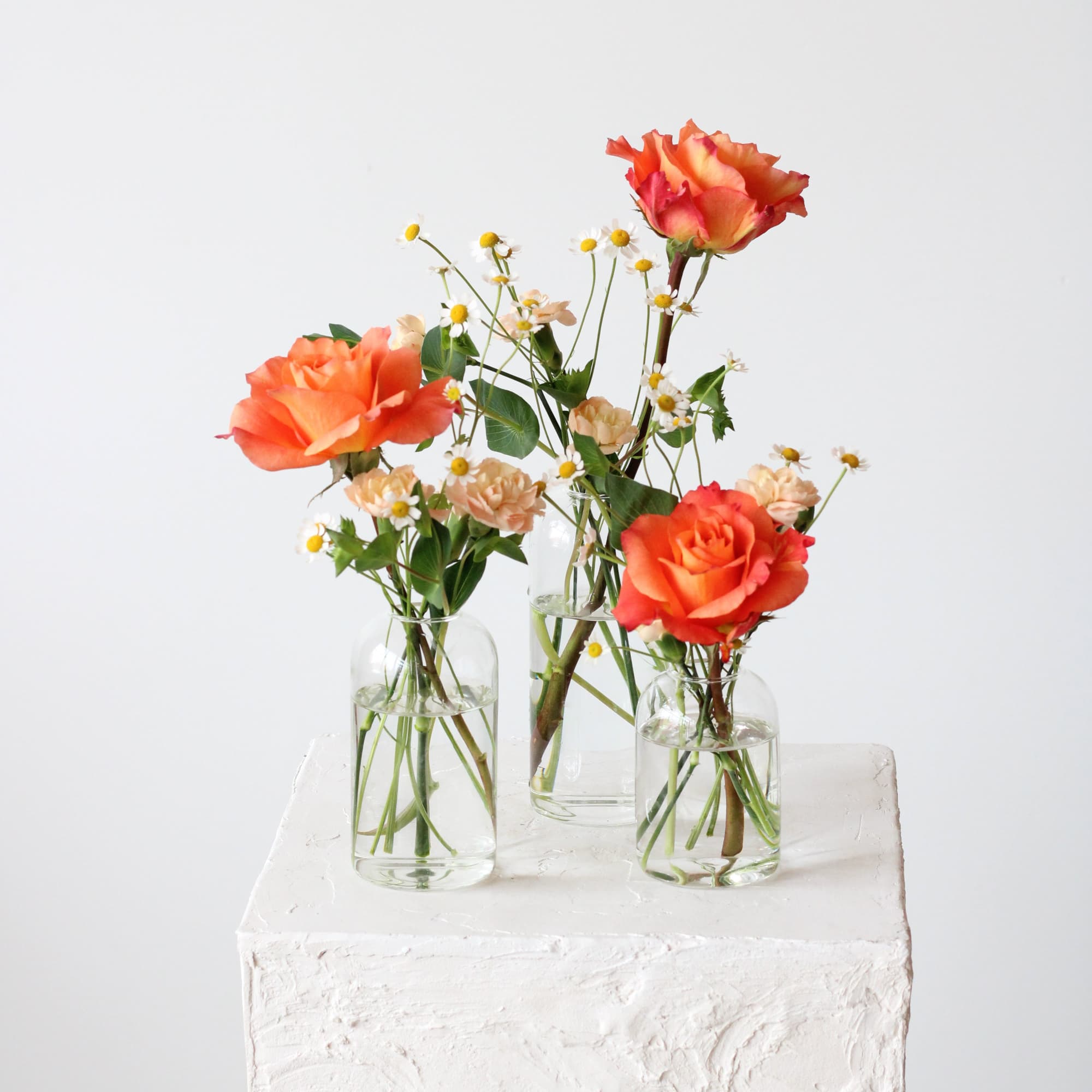 Orange roses and small white daisies in clear glass vases on a white pedestal.