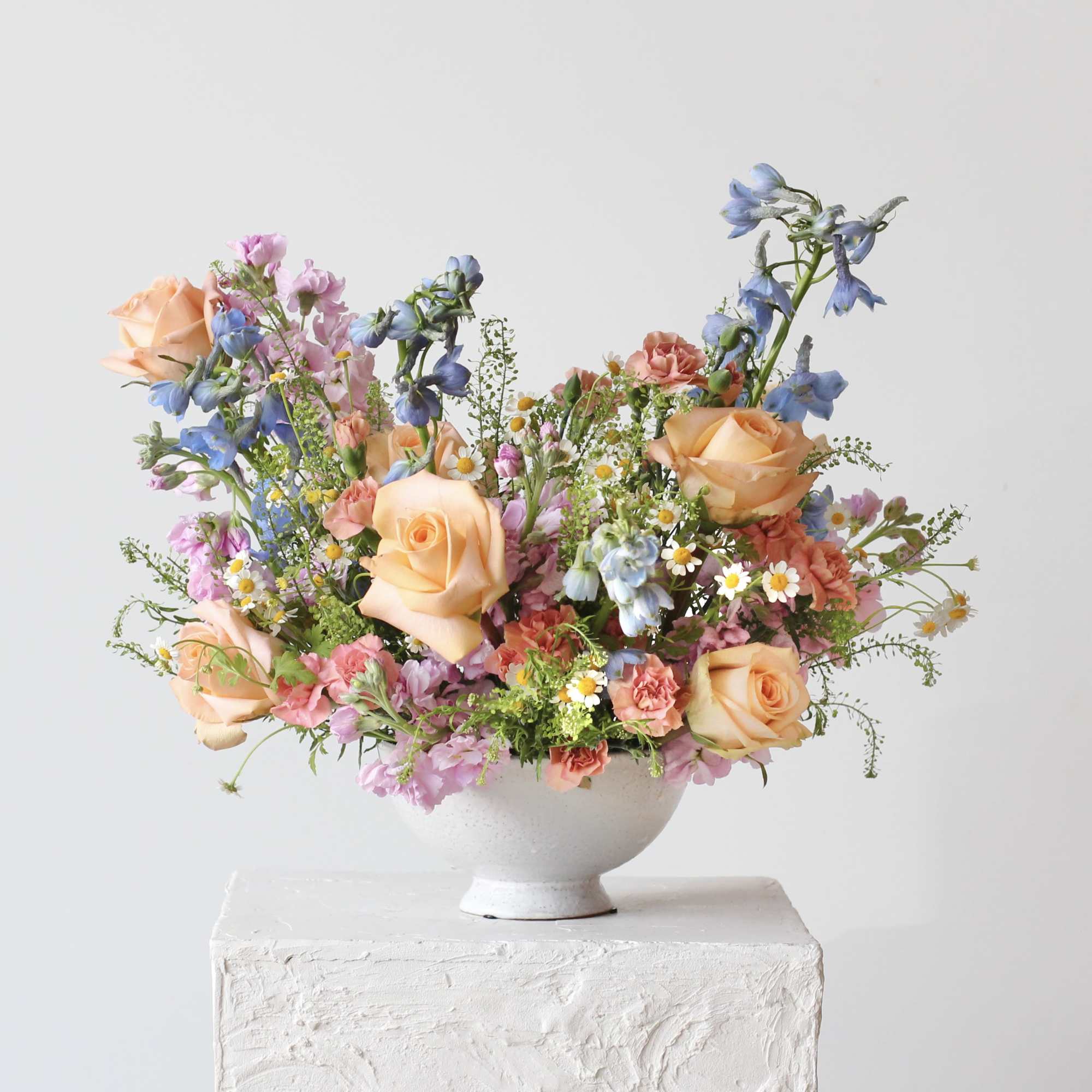 Mixed bouquet of peach roses, pink blooms, and blue flowers in a white bowl vase