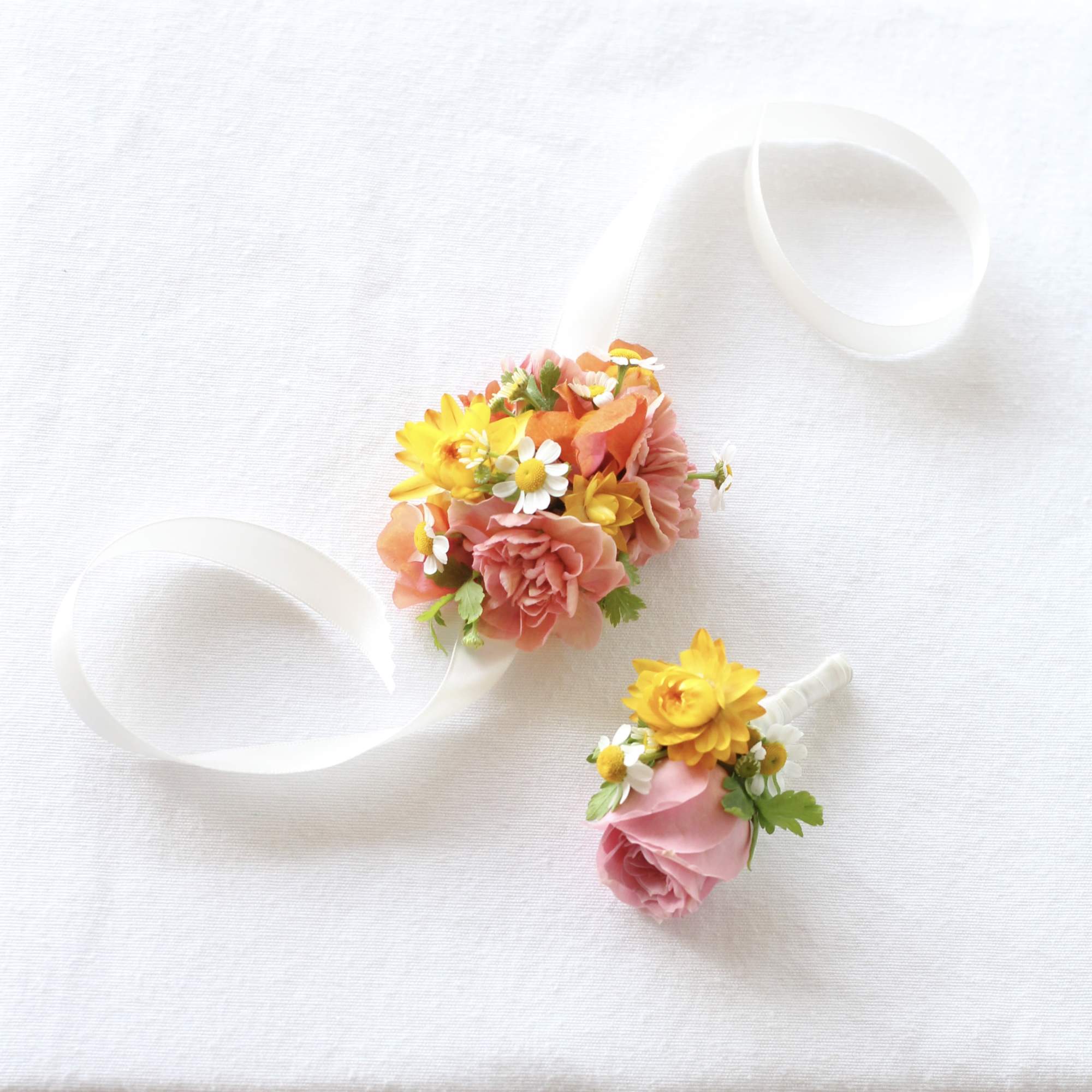 Two small floral boutonnieres with pink and yellow flowers on white ribbon