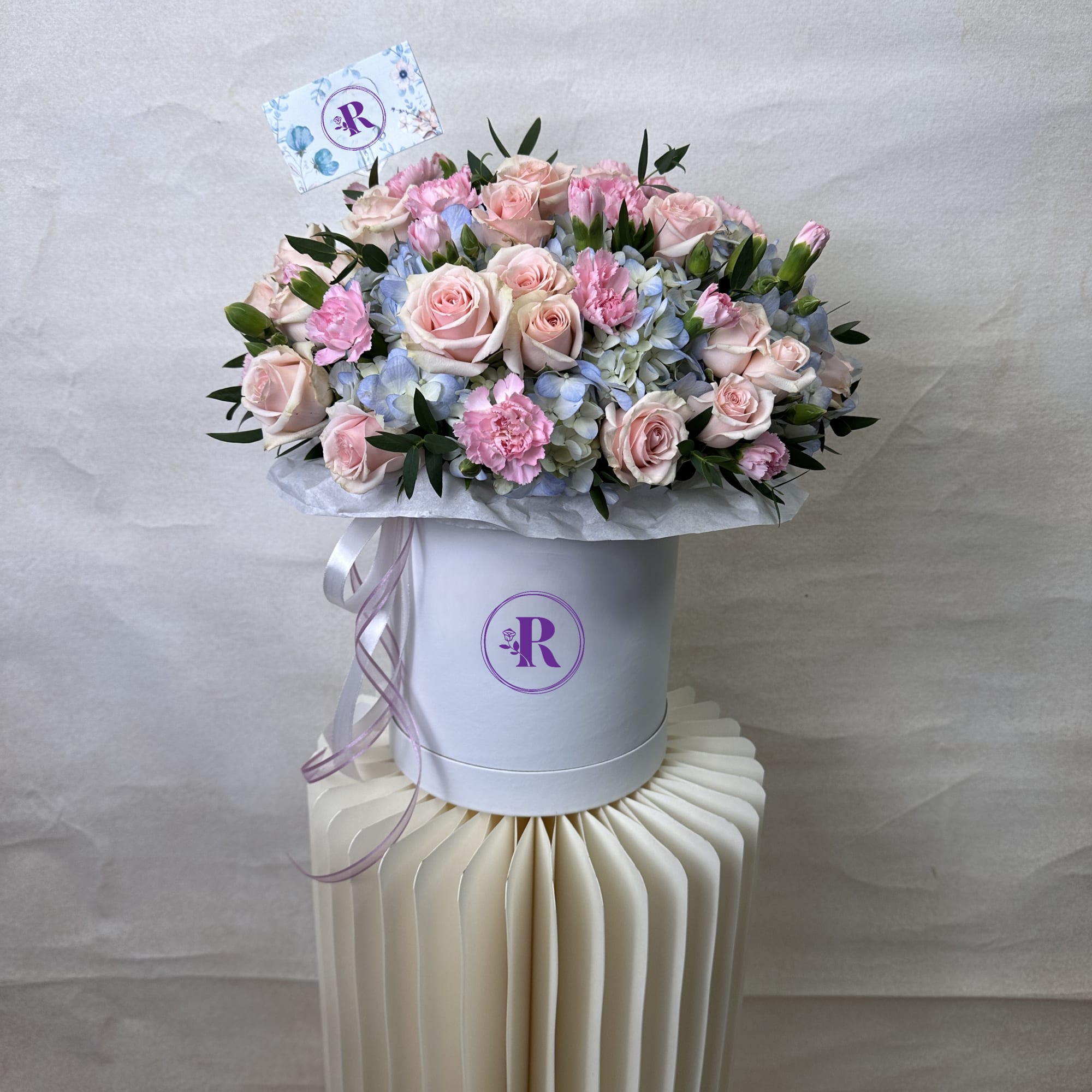 Round hatbox arrangement of pink roses, pink carnations, and blue hydrangeas