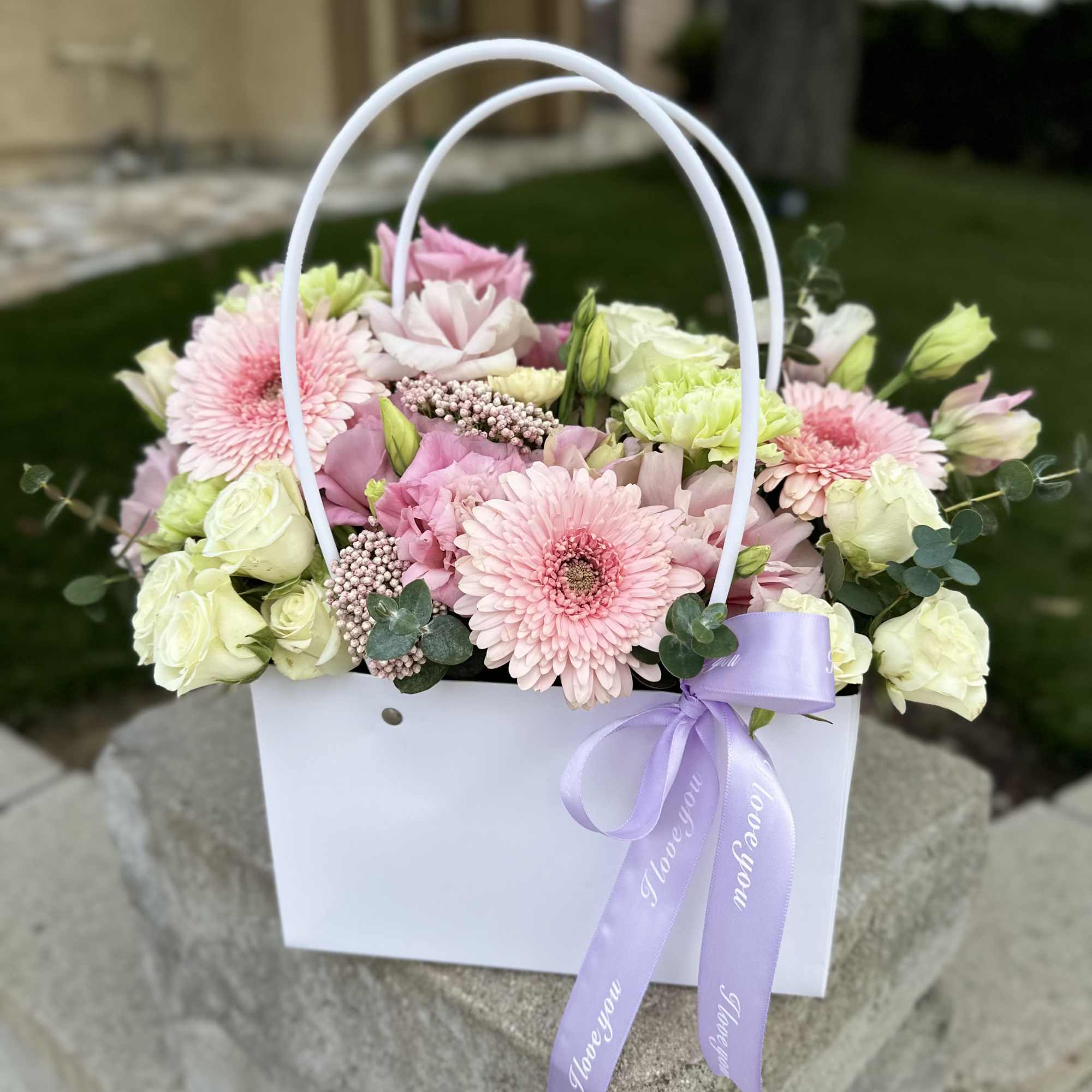 Pink and white flowers arranged in a white gift bag with a lavender ribbon