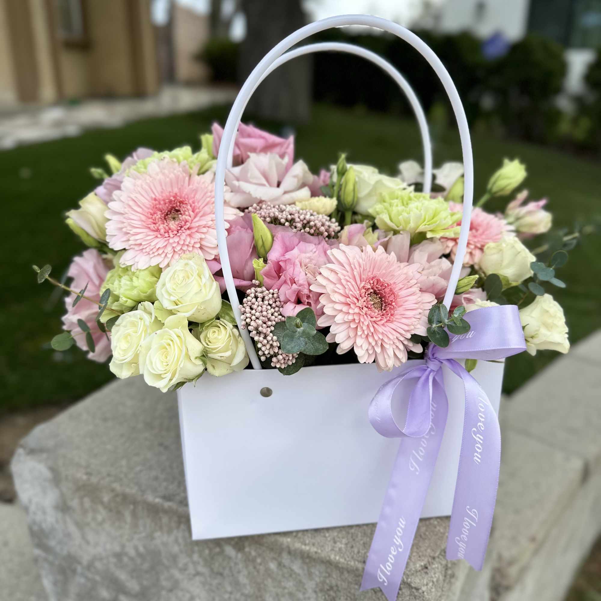 Pink and white flowers arranged in a white gift basket with a lavender ribbon