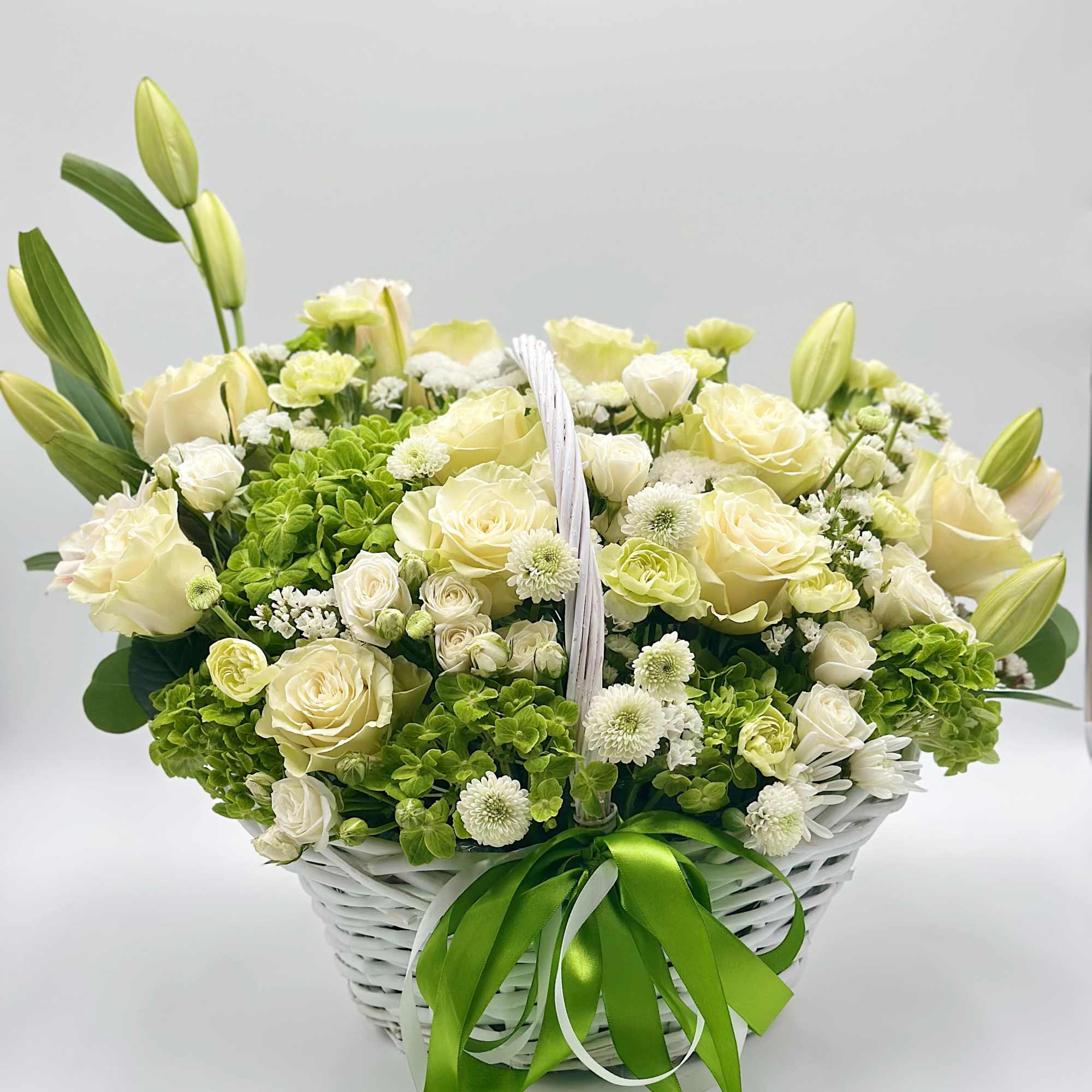 White basket arrangement of cream roses, lilies, and green hydrangeas with a green ribbon