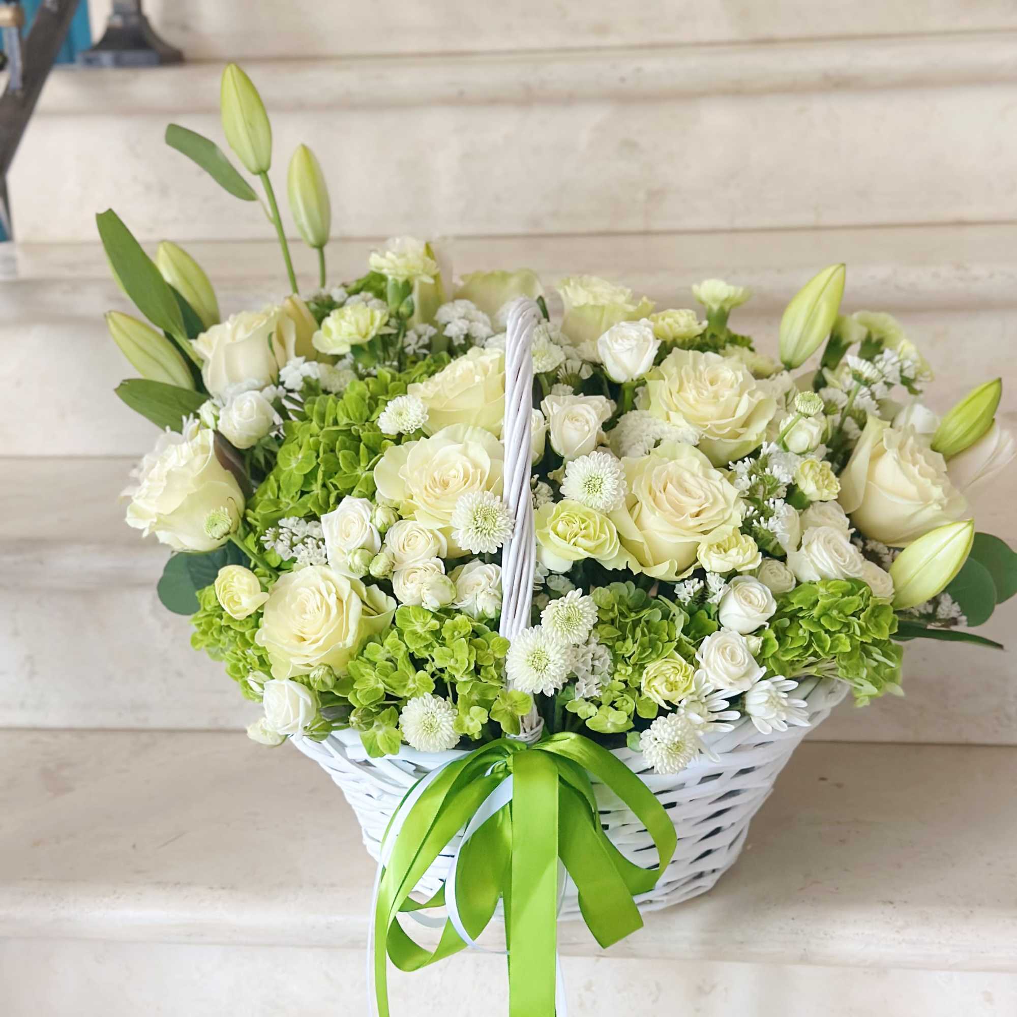 White basket of cream roses, lilies, and green hydrangeas with a green ribbon