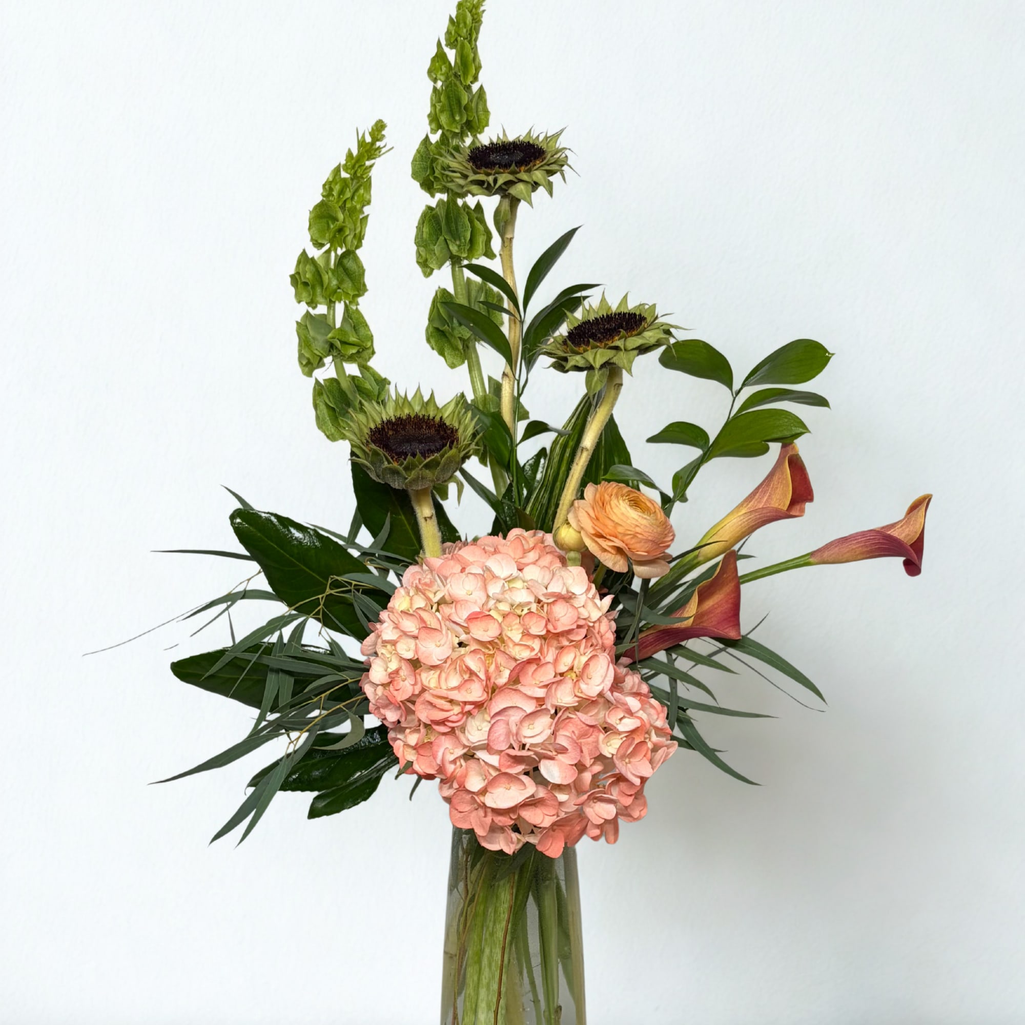 Tall floral arrangement with pink hydrangea, sunflowers, and calla lilies in a glass vase