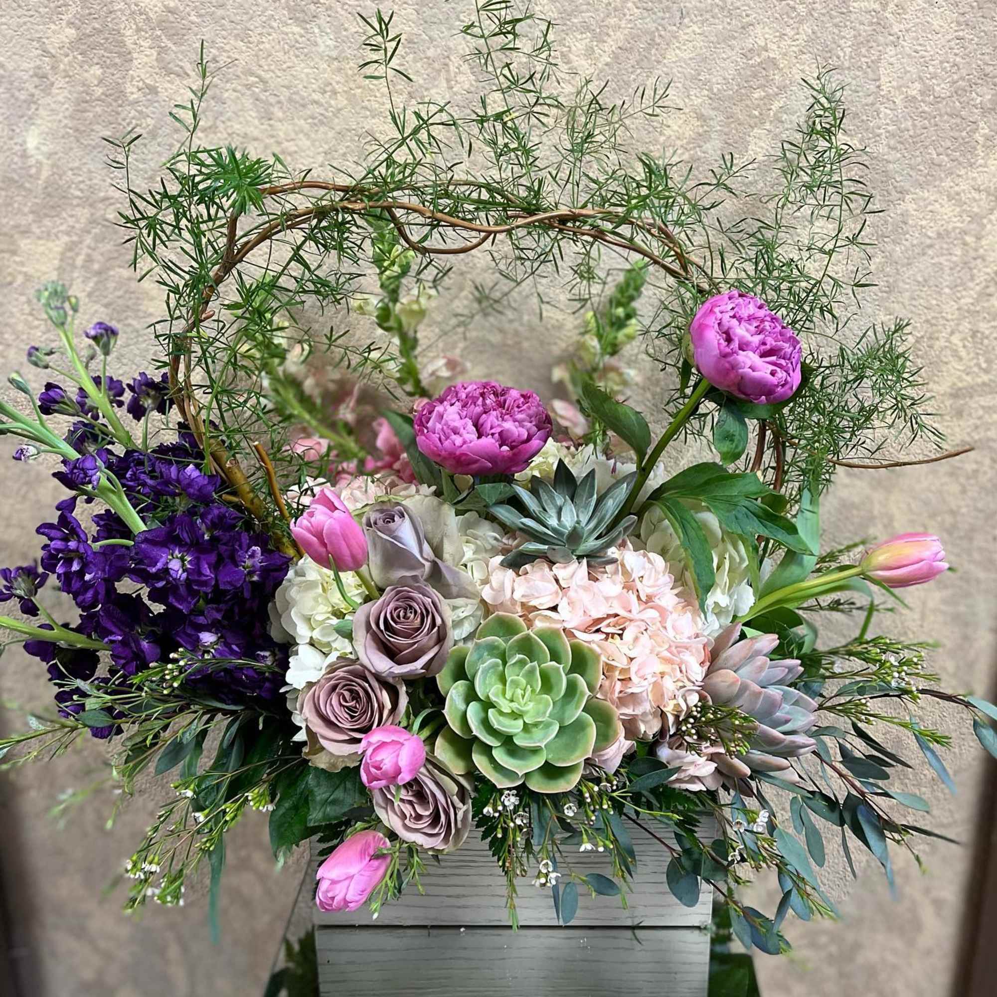 Lush arrangement of pink peonies, roses, tulips, succulents and hydrangeas in a gray wooden box with a vine hoop.