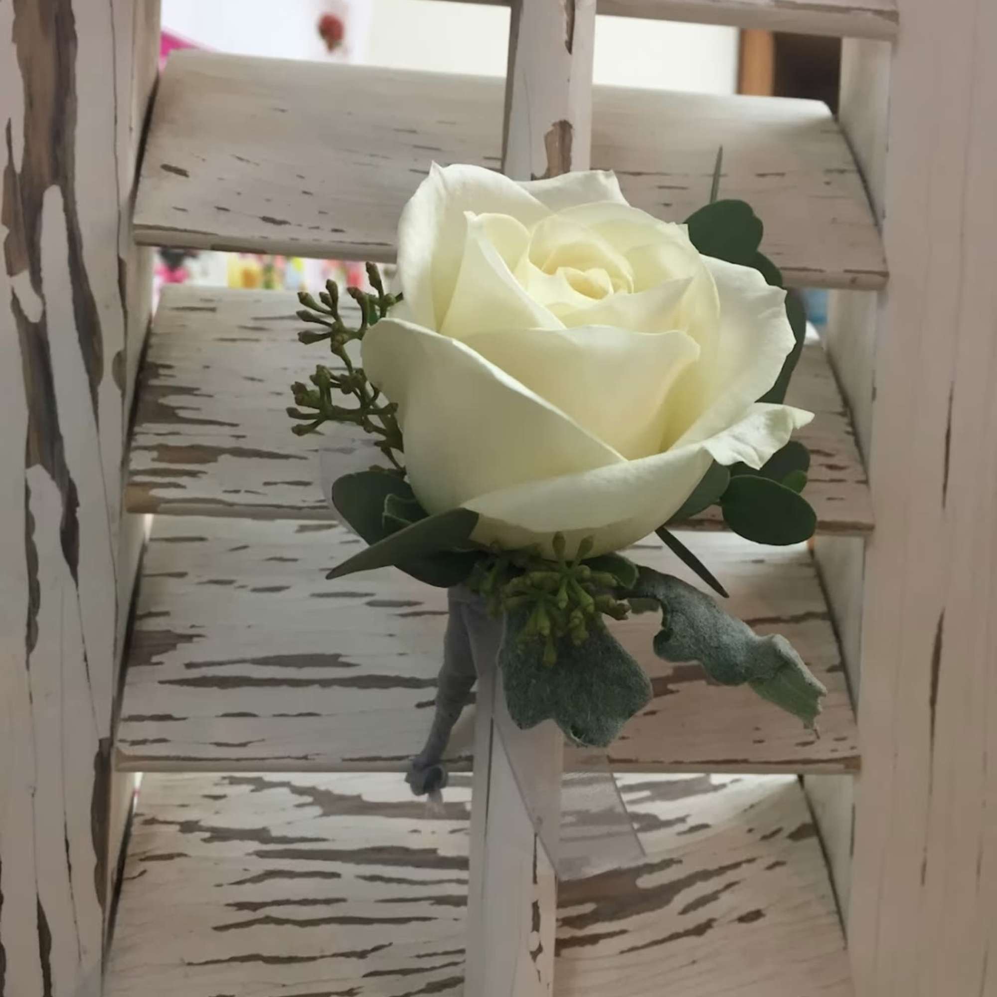 Single white rose boutonniere with greenery and ribbon on a rustic white wooden shutter