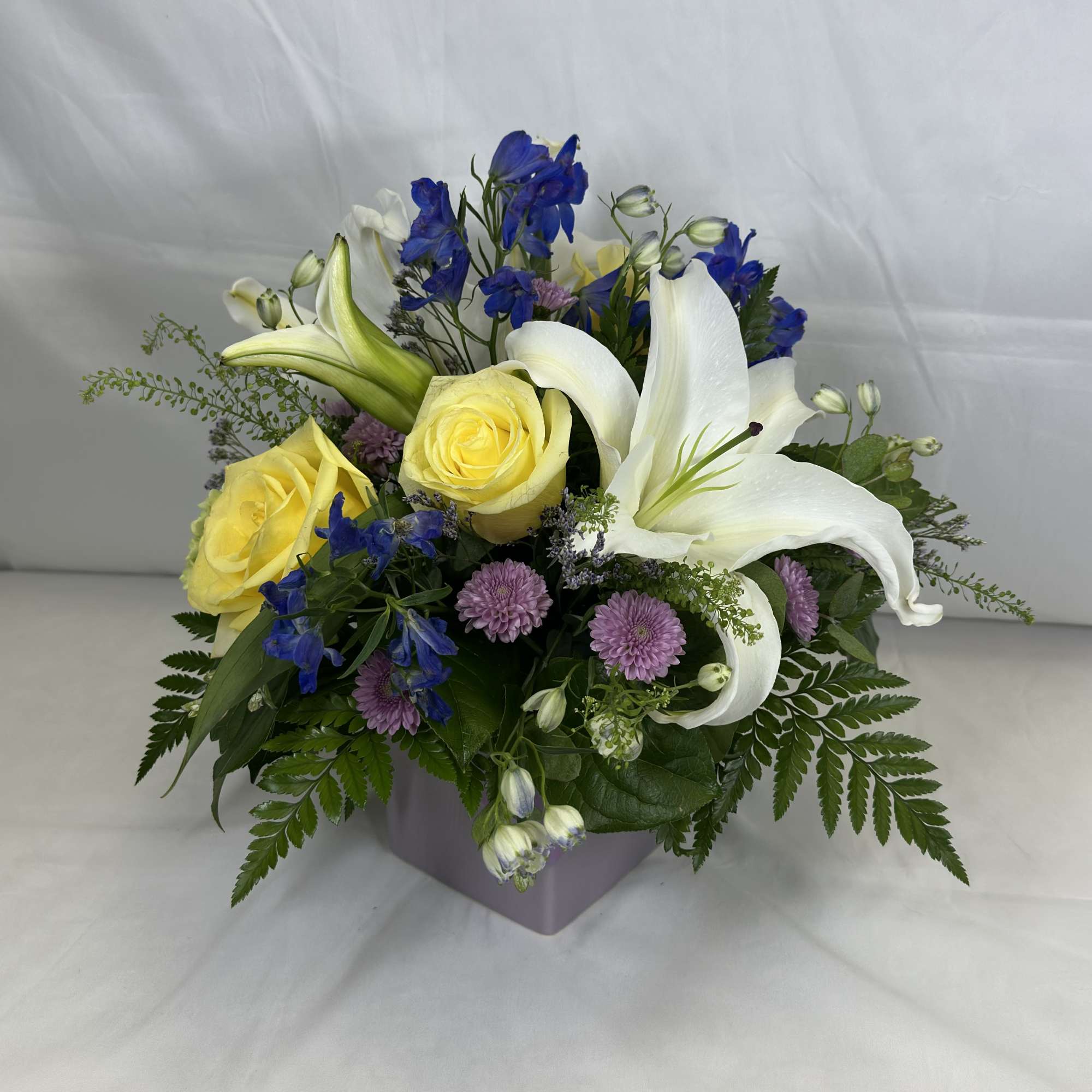 Bouquet of yellow roses, white lilies, and blue flowers in a square vase