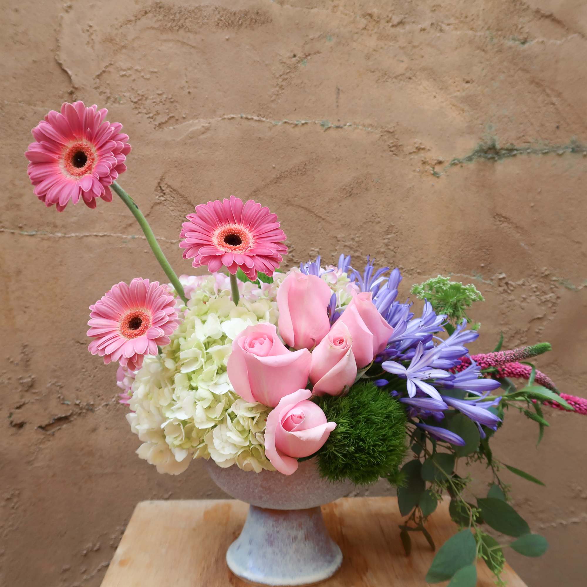 Pink gerberas and roses arranged in a pedestal vase with purple flowers
