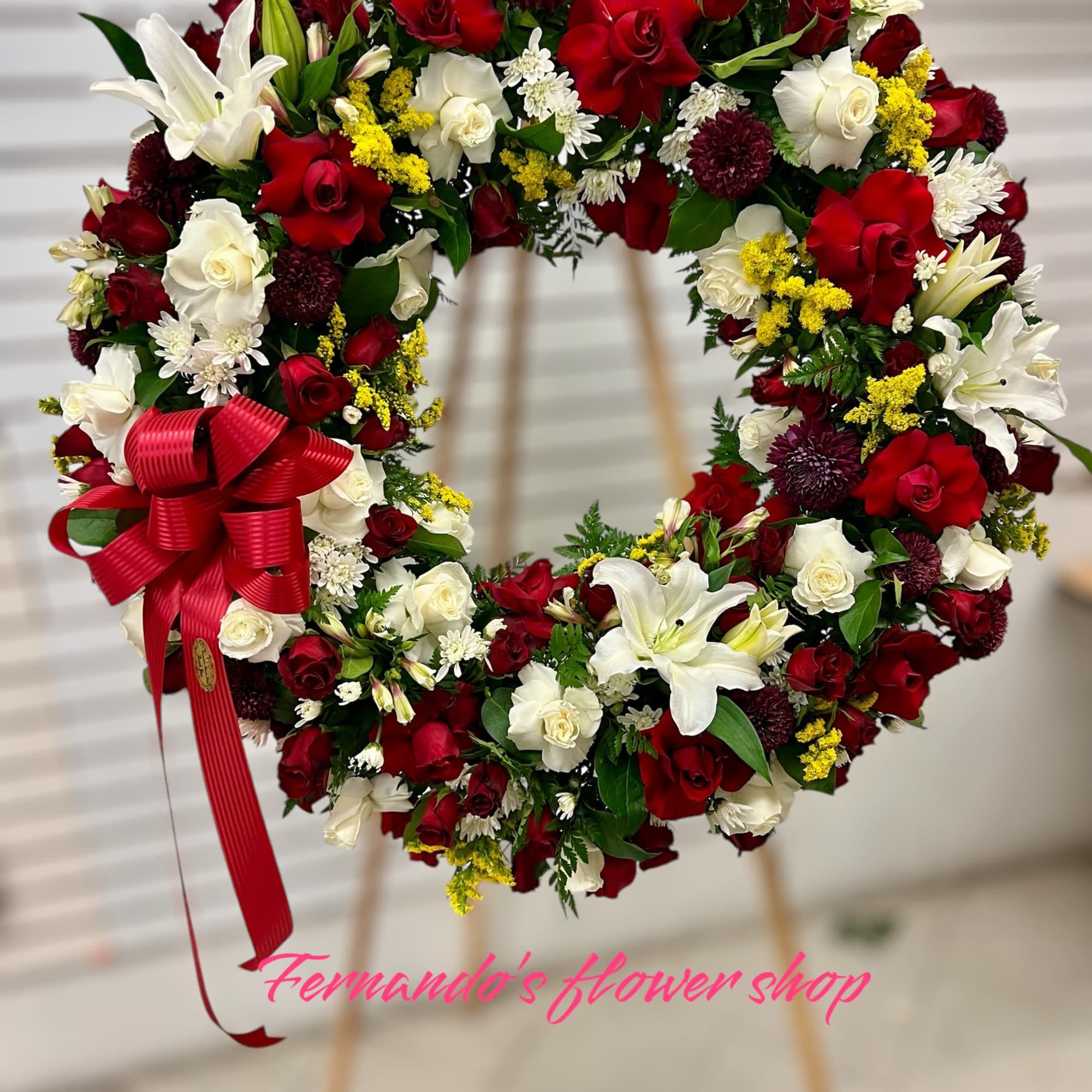 Standing wreath of red and white roses with white lilies, mums, and a red bow on an easel.