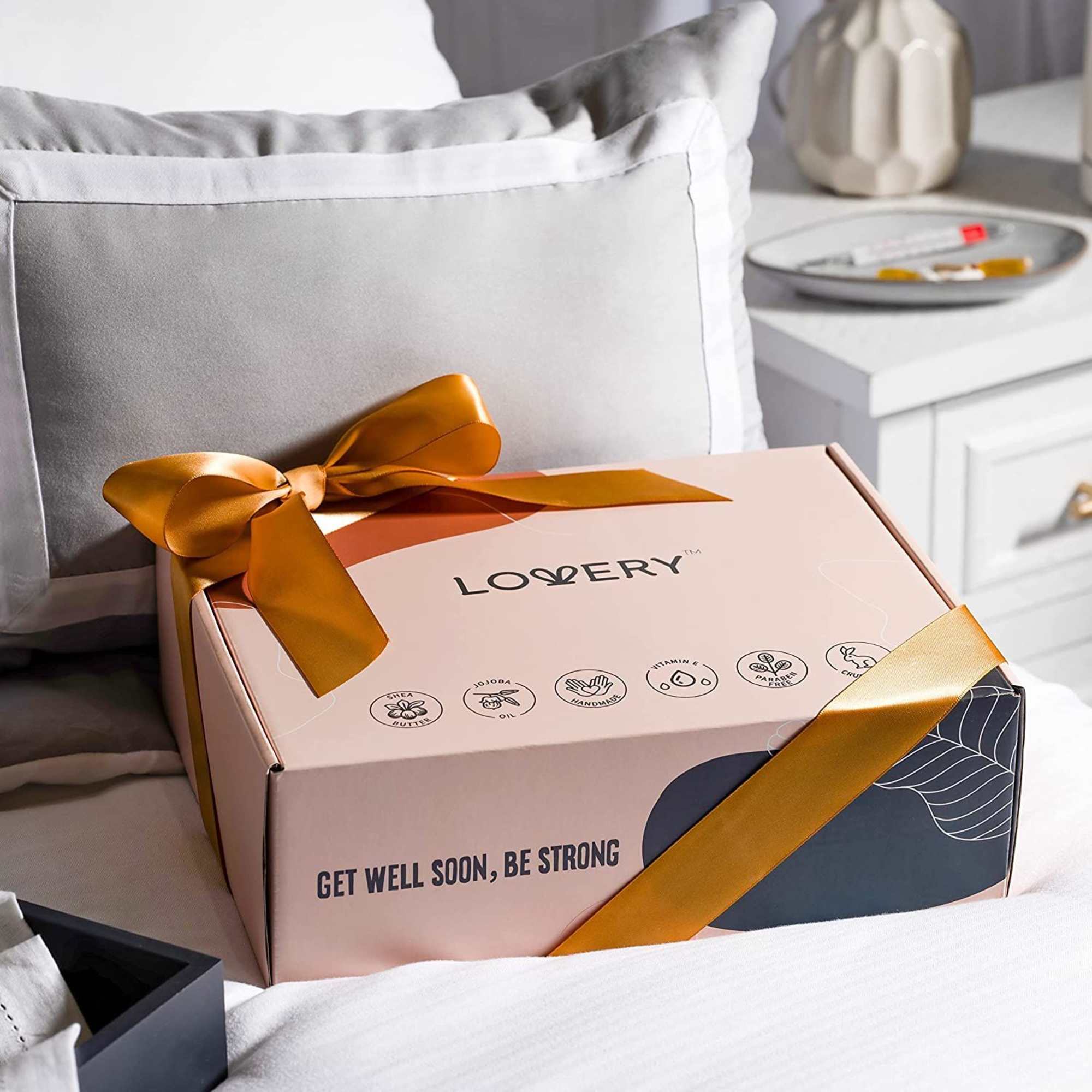 Peach gift box with gold ribbon reading 'Get Well Soon, Be Strong' resting on a neatly made bed