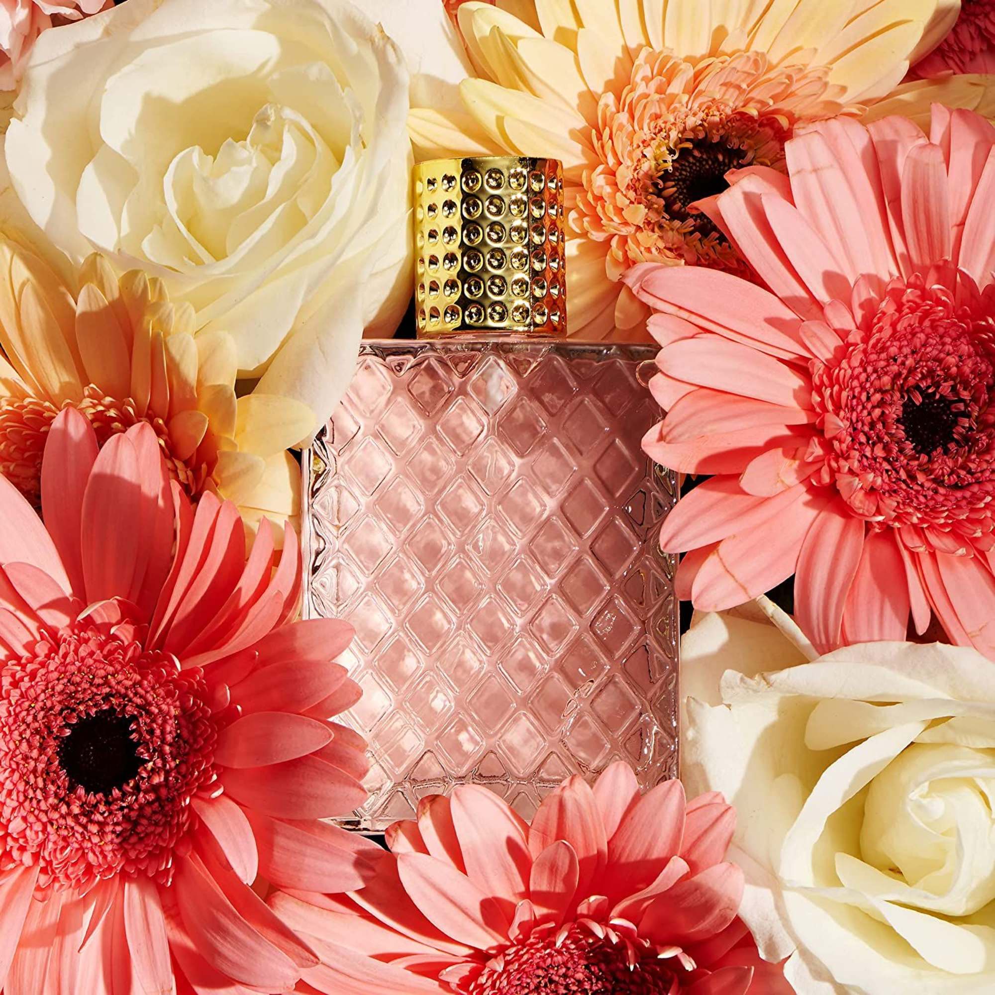 Pink perfume bottle with gold cap surrounded by white roses and pink gerbera daisies