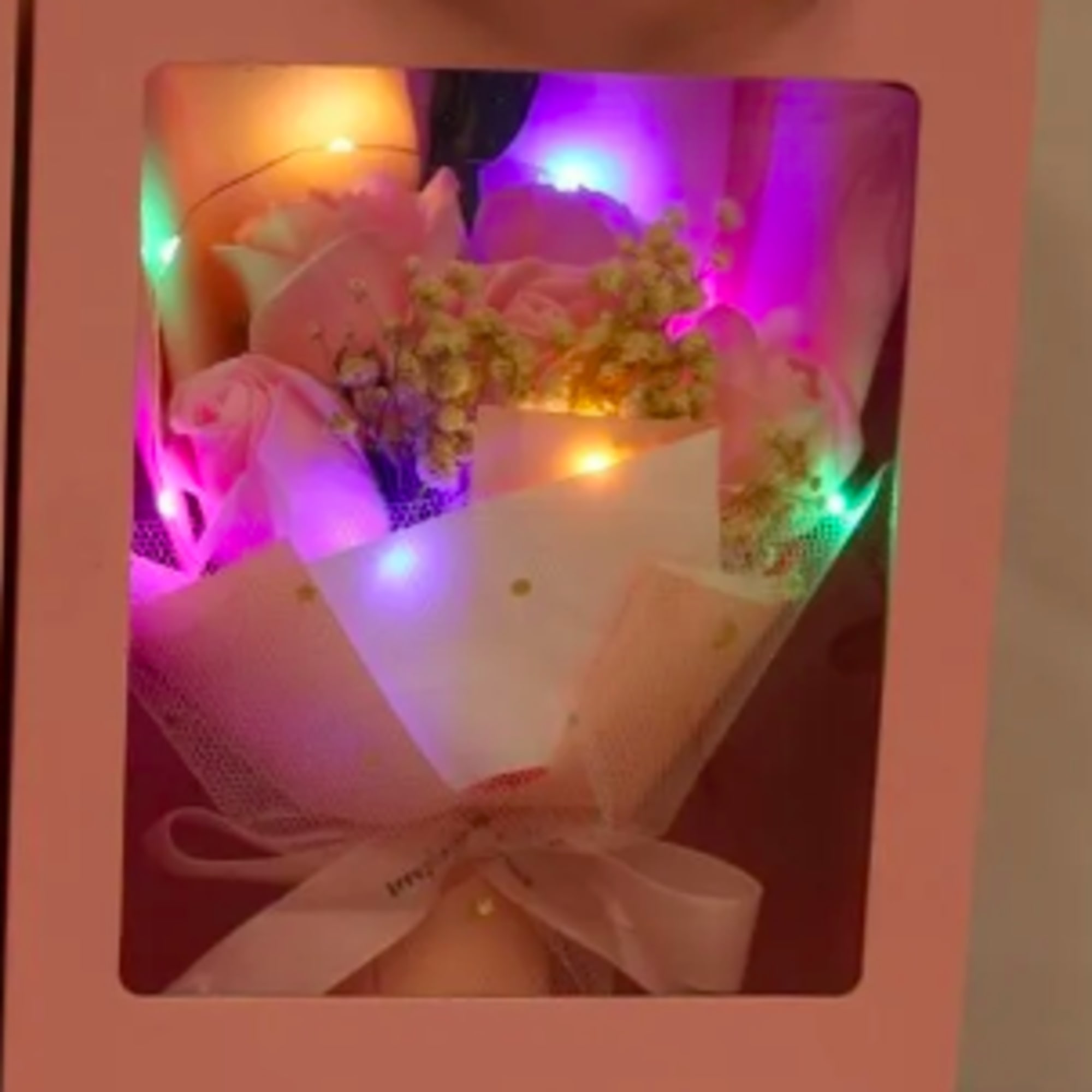 Pink gift box displaying a bouquet of pink roses with filler flowers and colorful string lights.