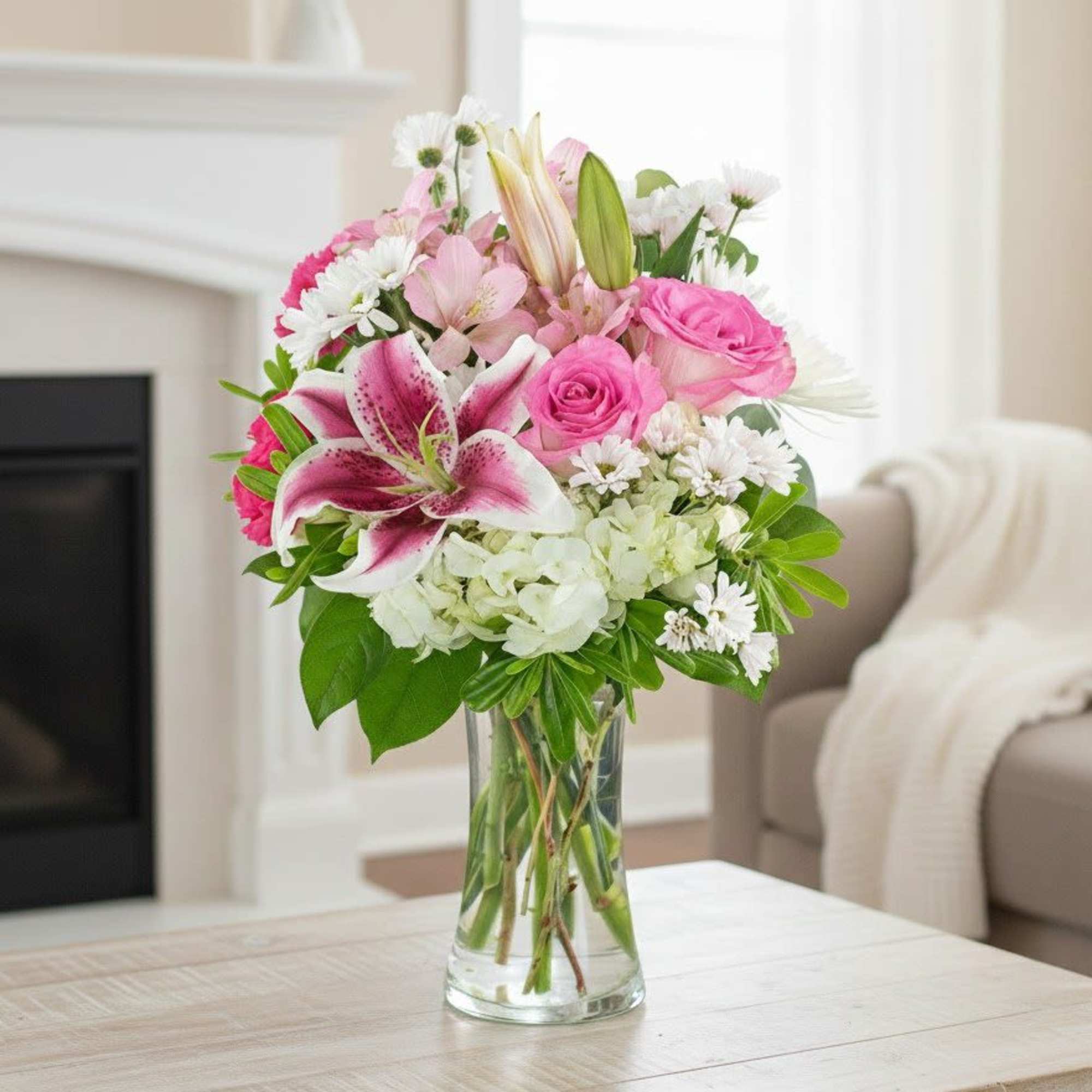 Pink lilies, roses, and white blooms arranged in a clear glass vase on a light wood table