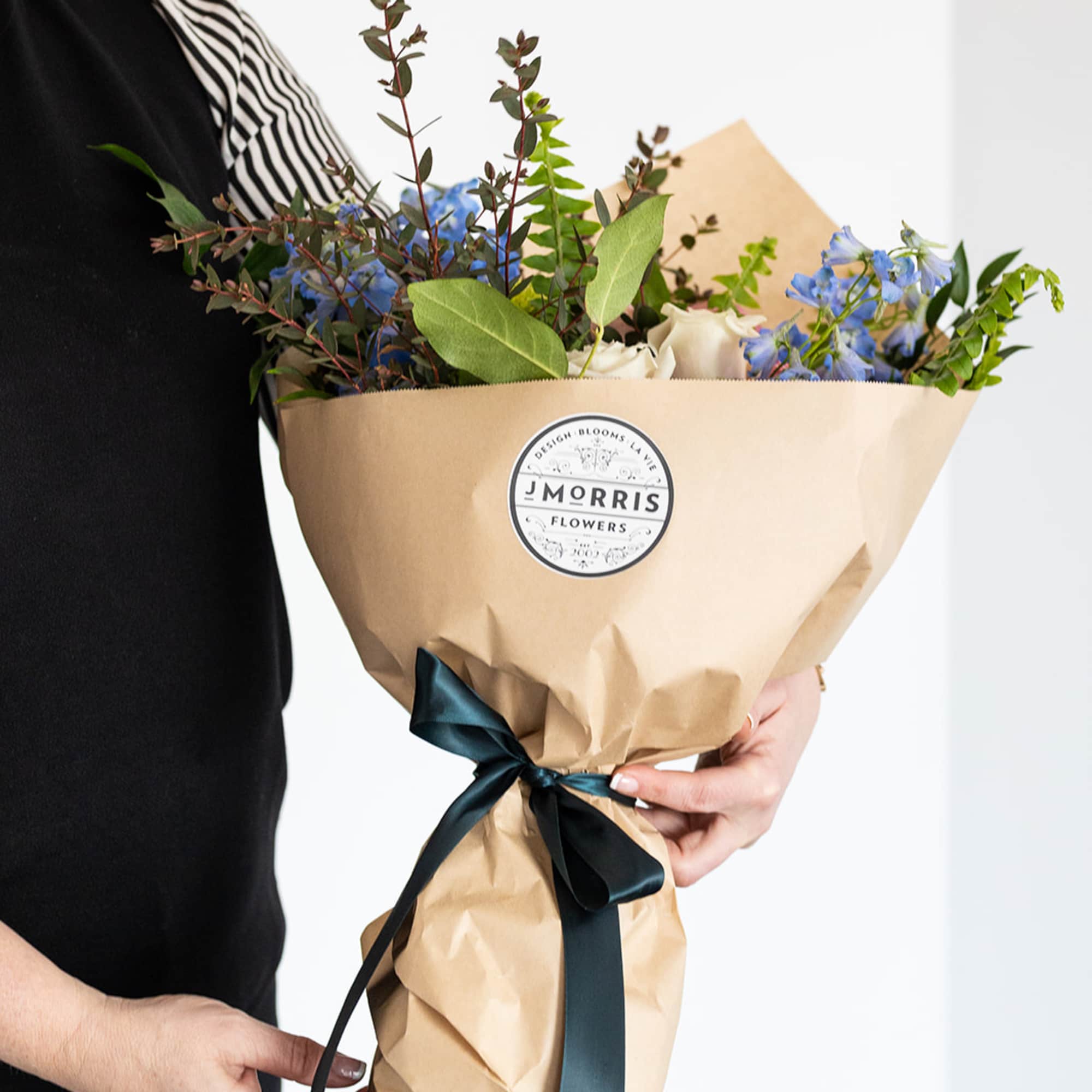 Hand-tied bouquet of blue and white flowers in kraft paper with a dark ribbon