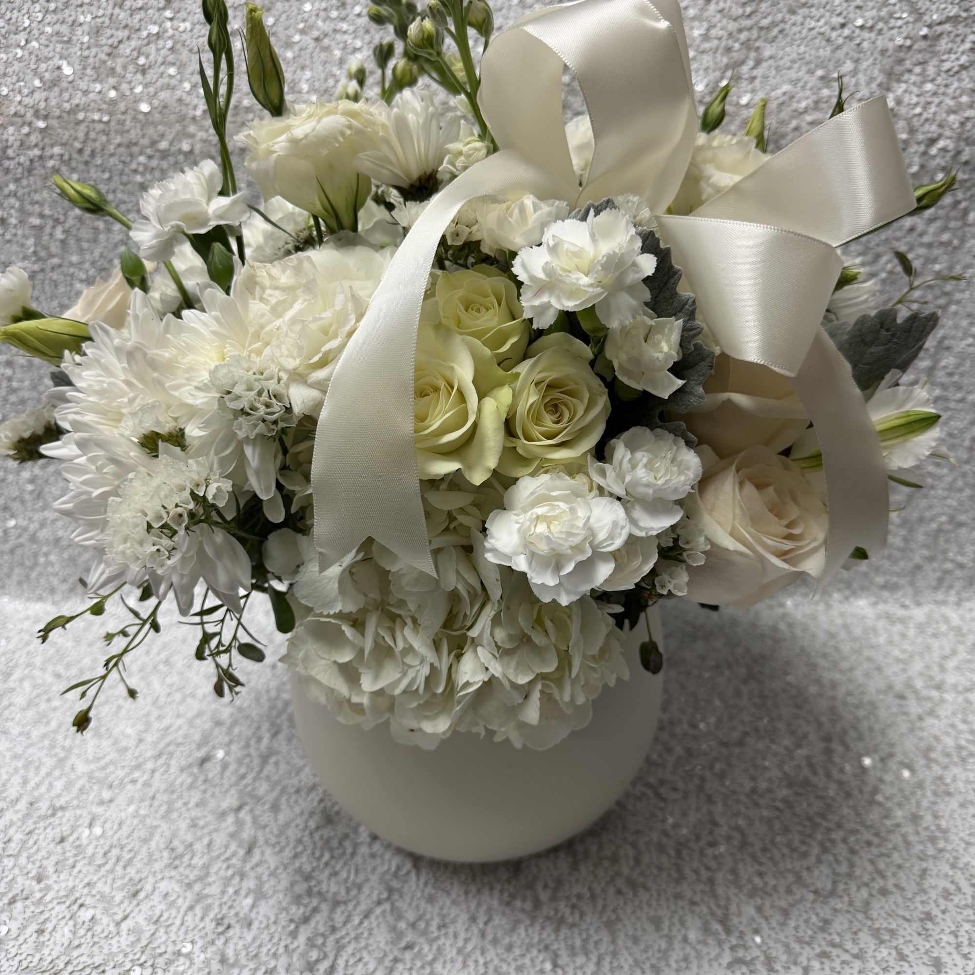 White floral arrangement with roses, carnations, and a satin ribbon in a vase