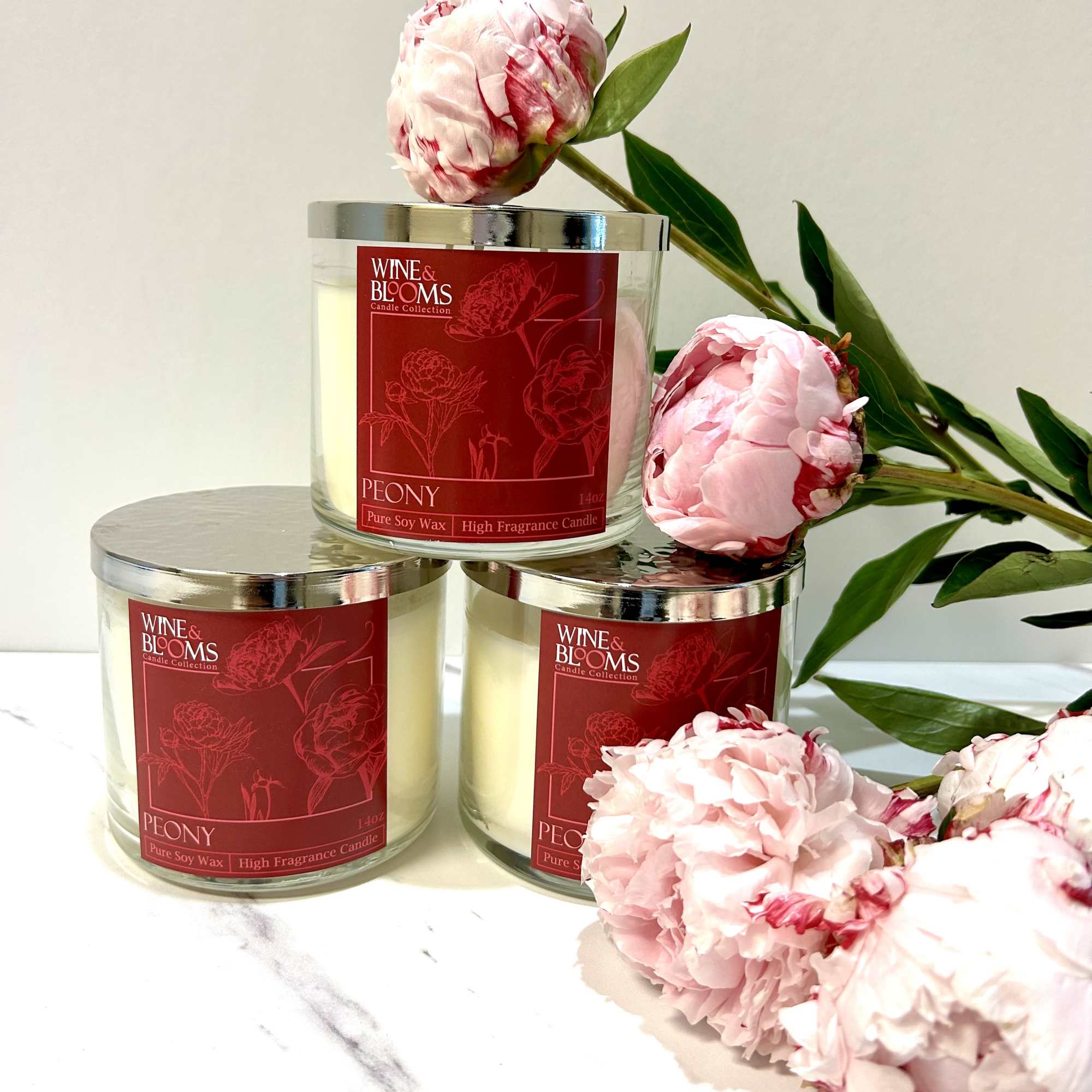 Three peony-scented candles with pink peony blooms arranged around them
