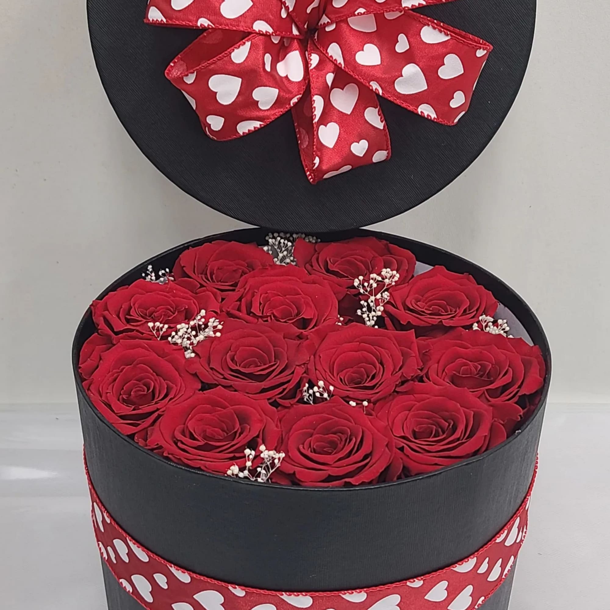 Round black hatbox filled with red roses and white filler flowers, trimmed with red heart-print ribbon.
