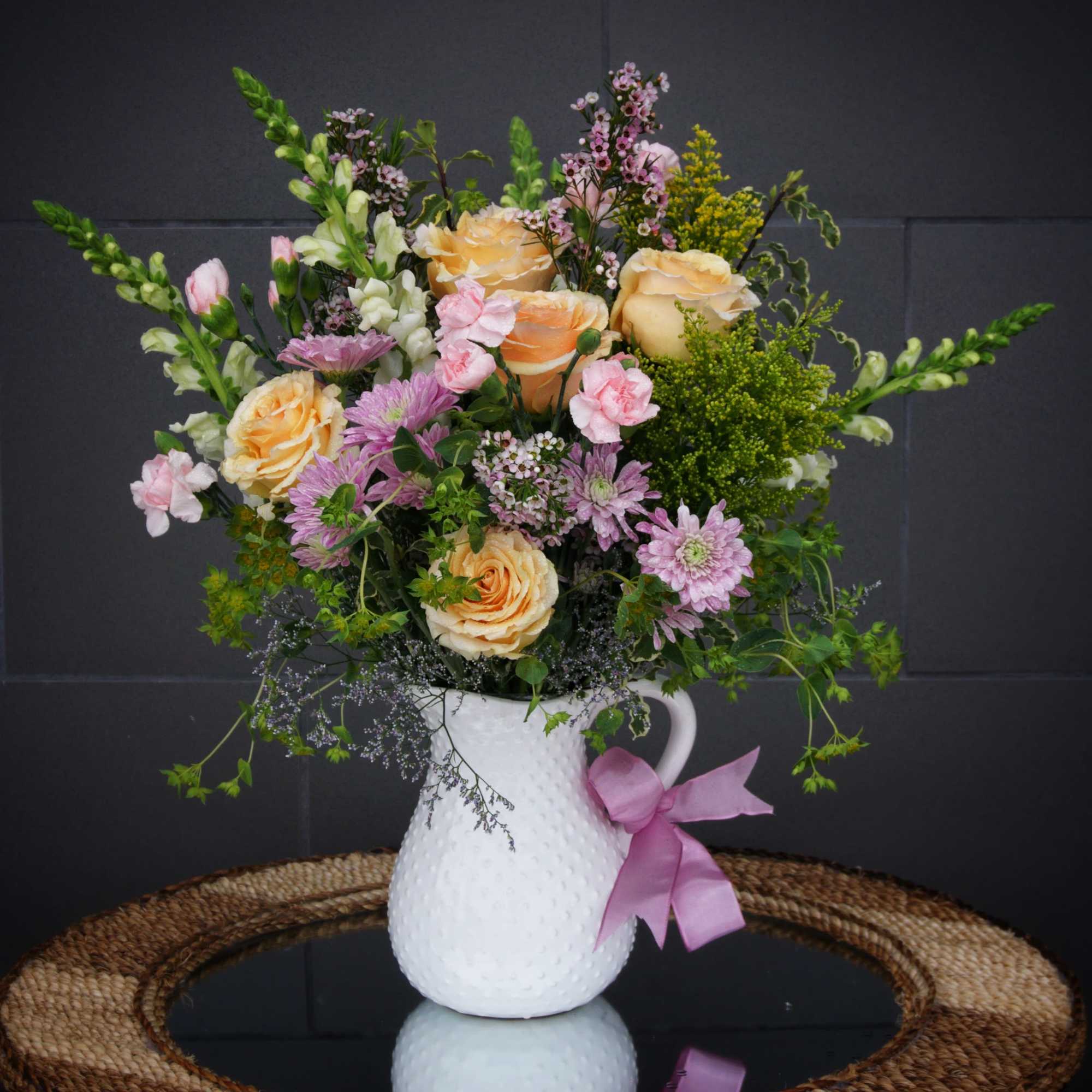 Peach roses and pink flowers in a white pitcher vase with a pink ribbon