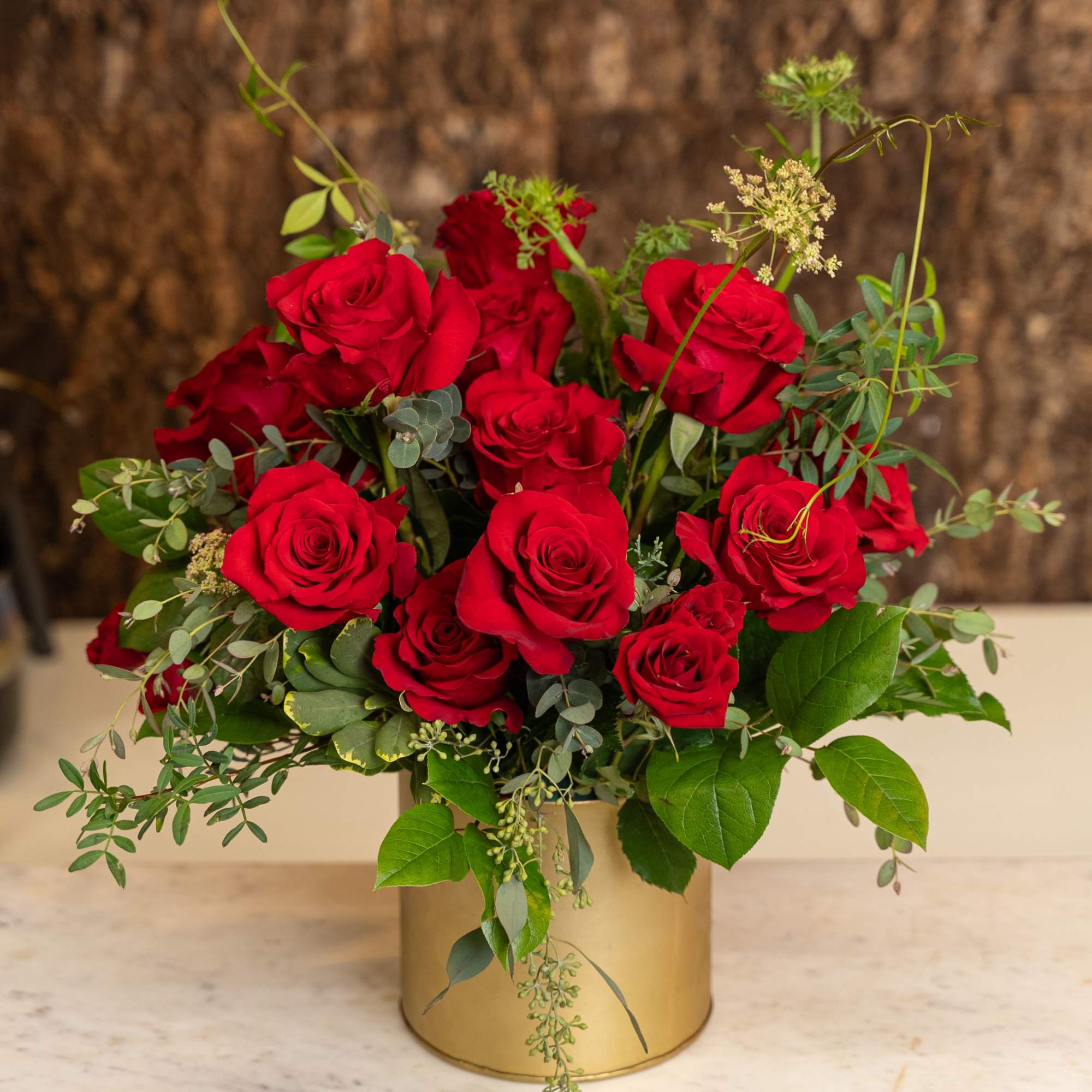 Round arrangement of red roses with assorted foliage in a gold cylinder vase
