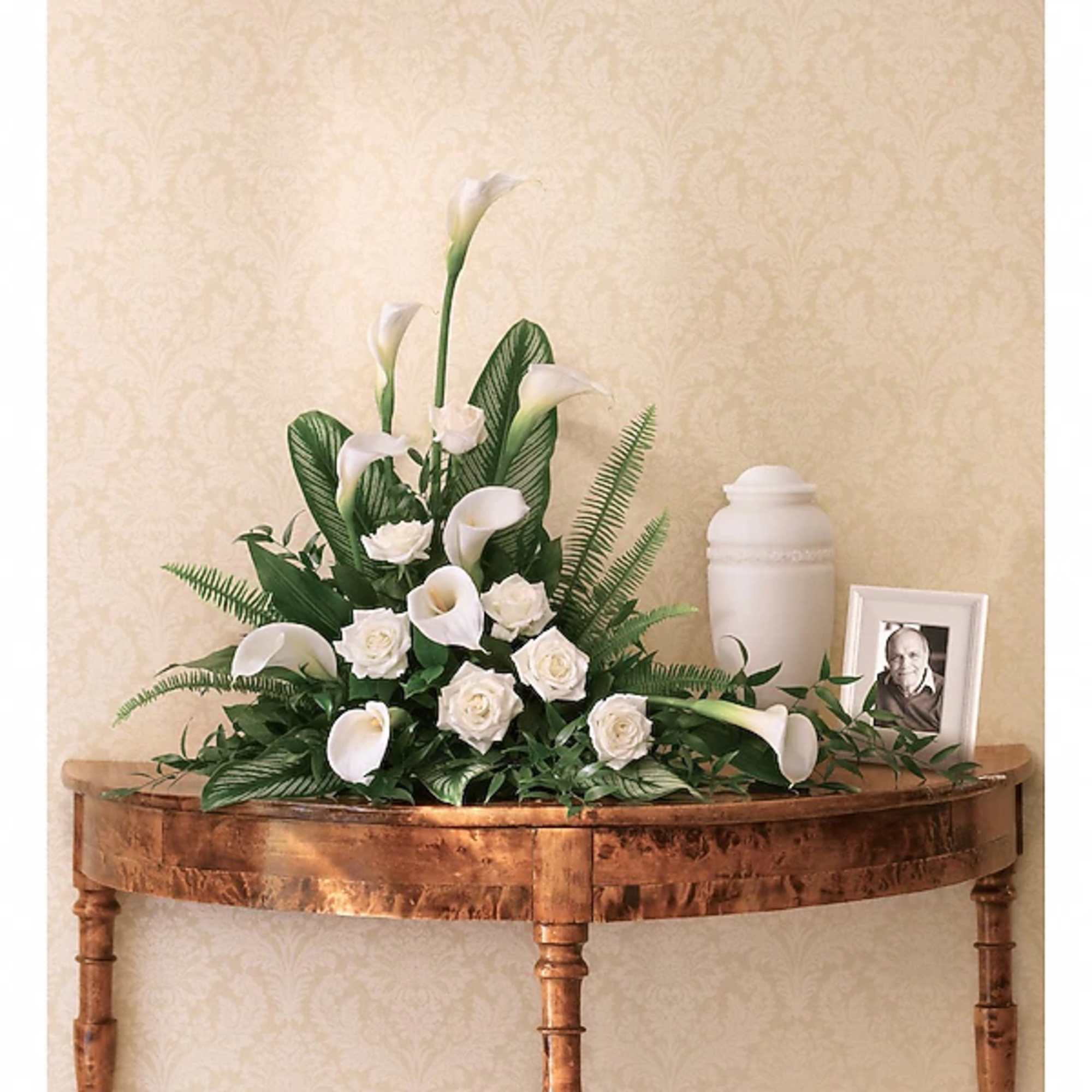 White calla lilies and roses arranged on a wooden table with a memorial urn and framed photo