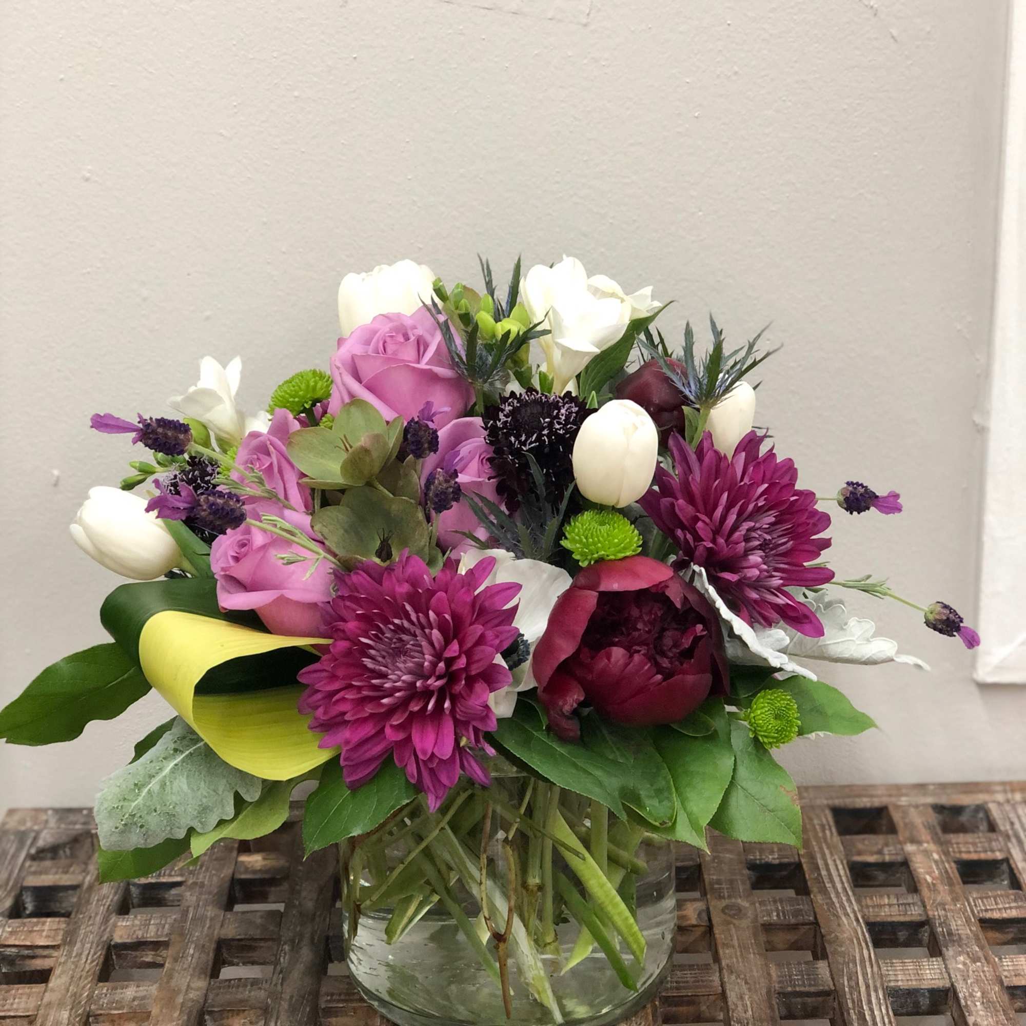 Low glass vase arrangement with purple mums, lavender roses, white tulips, and mixed accent blooms.