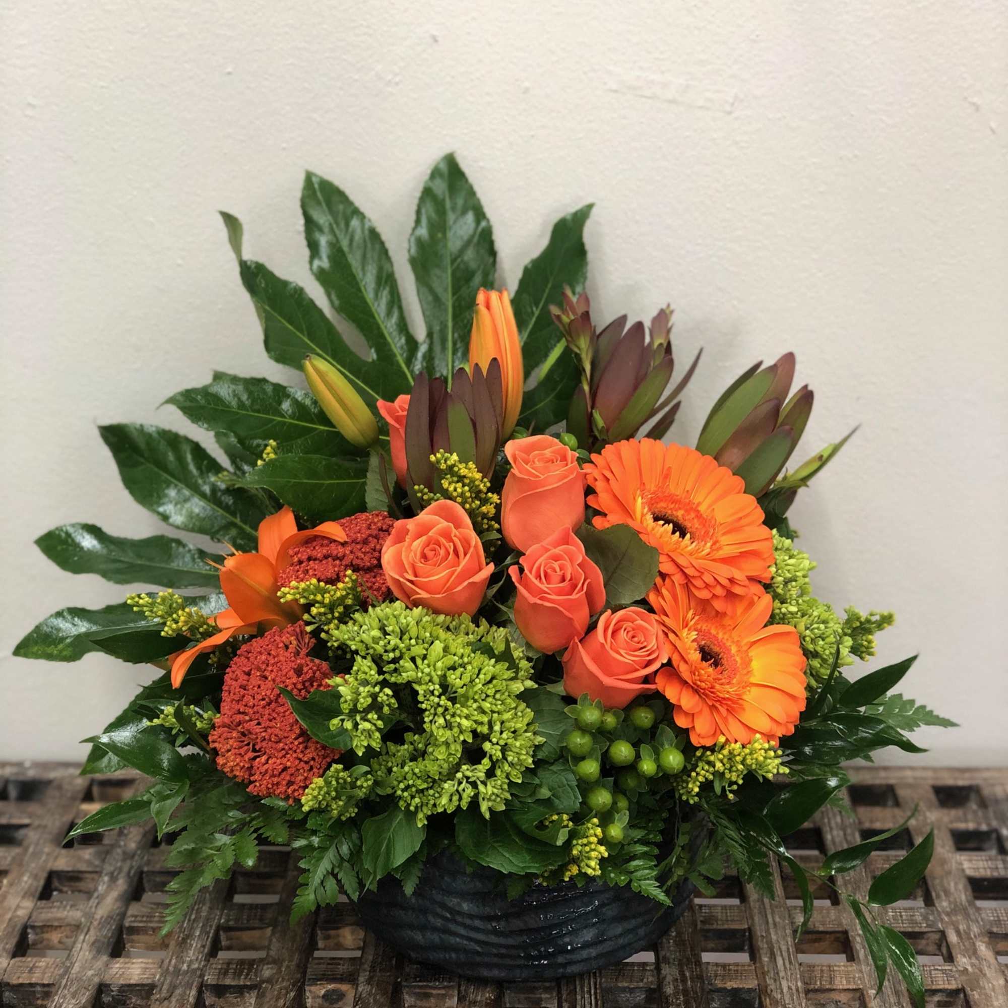 Low orange arrangement with roses, gerbera daisies, lilies and green accents in a black bowl