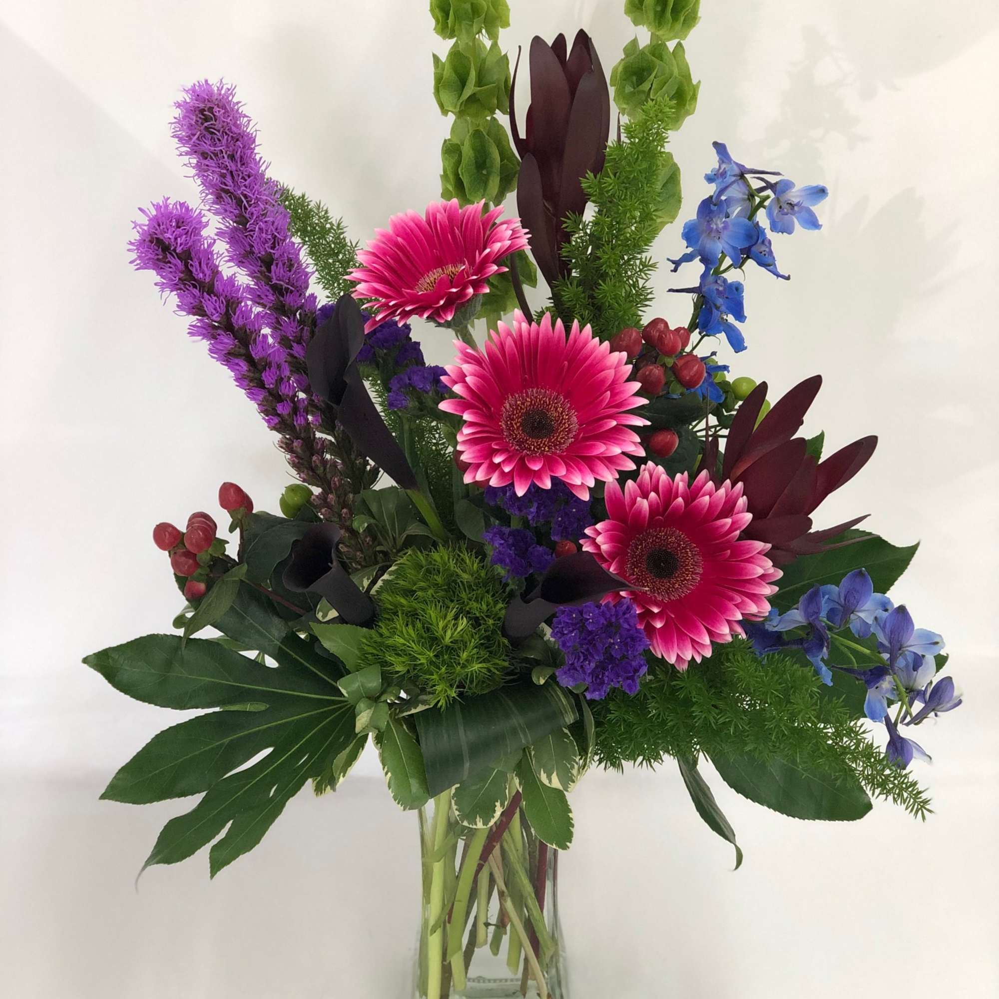 Tall mixed arrangement with pink gerbera daisies, purple and blue blooms, and green accents in a clear glass vase