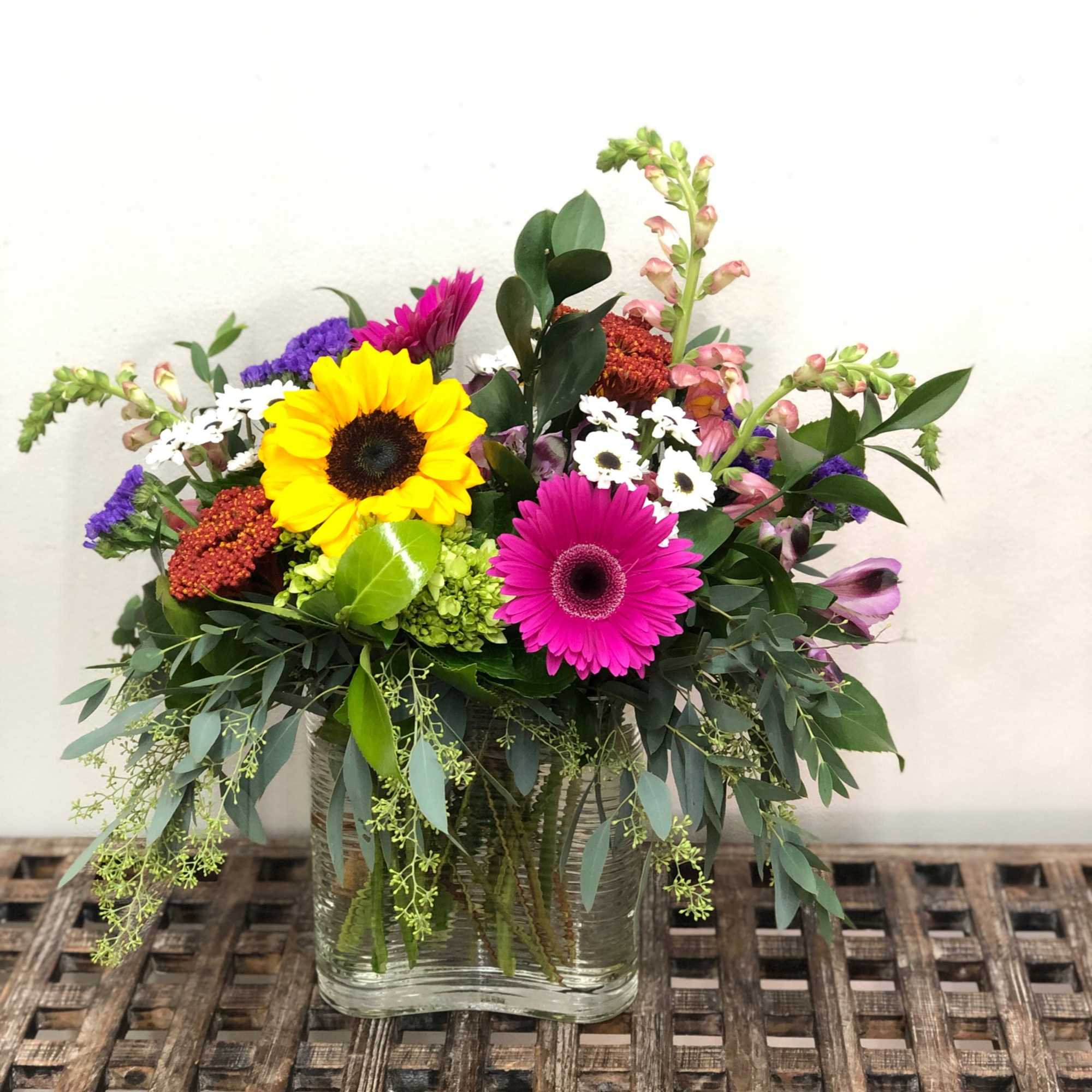 Colorful mixed arrangement with sunflower, pink gerbera daisy, and other blooms in a clear glass cube vase.