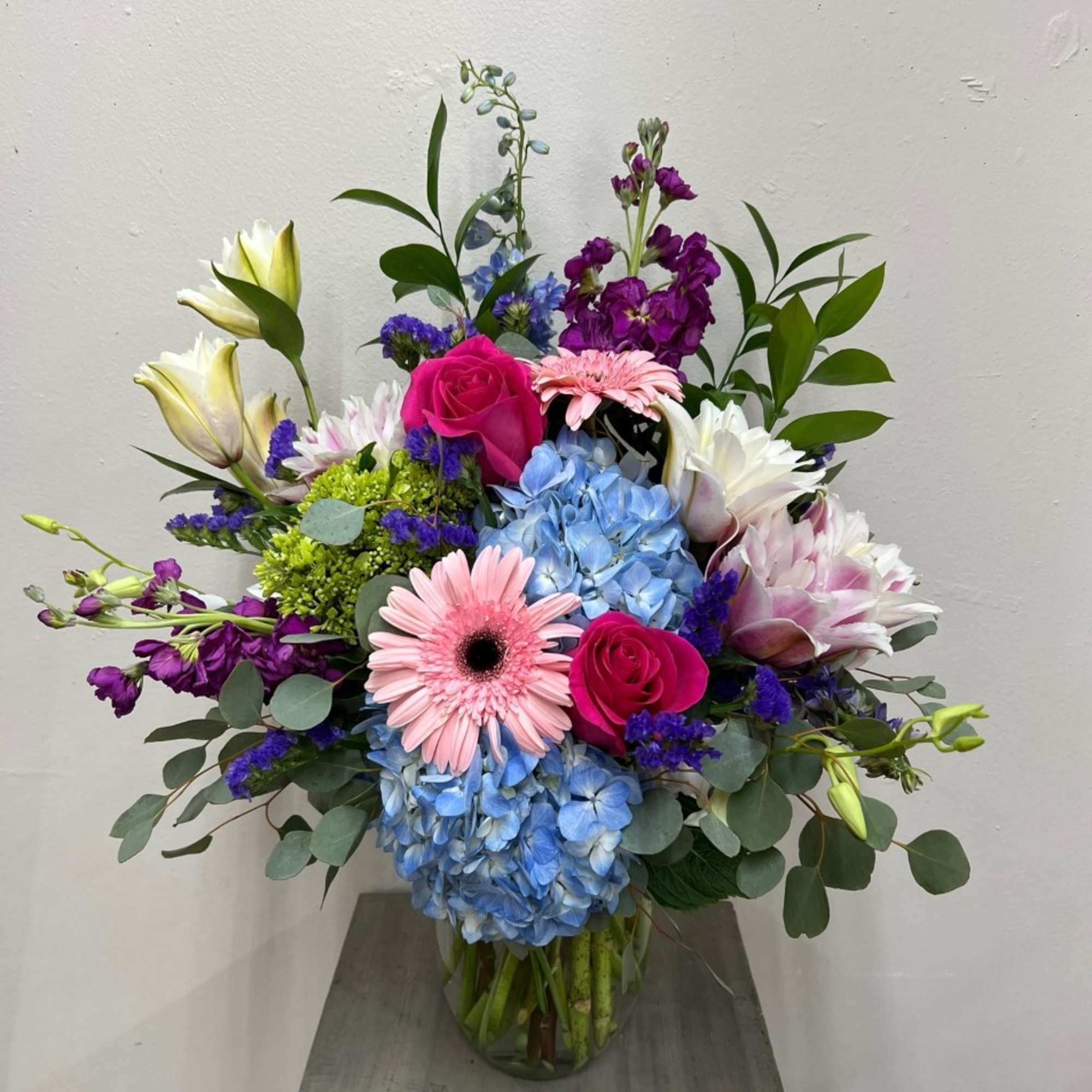 Mixed bouquet of blue hydrangeas, pink gerberas, roses, lilies and purple blooms in a clear glass vase