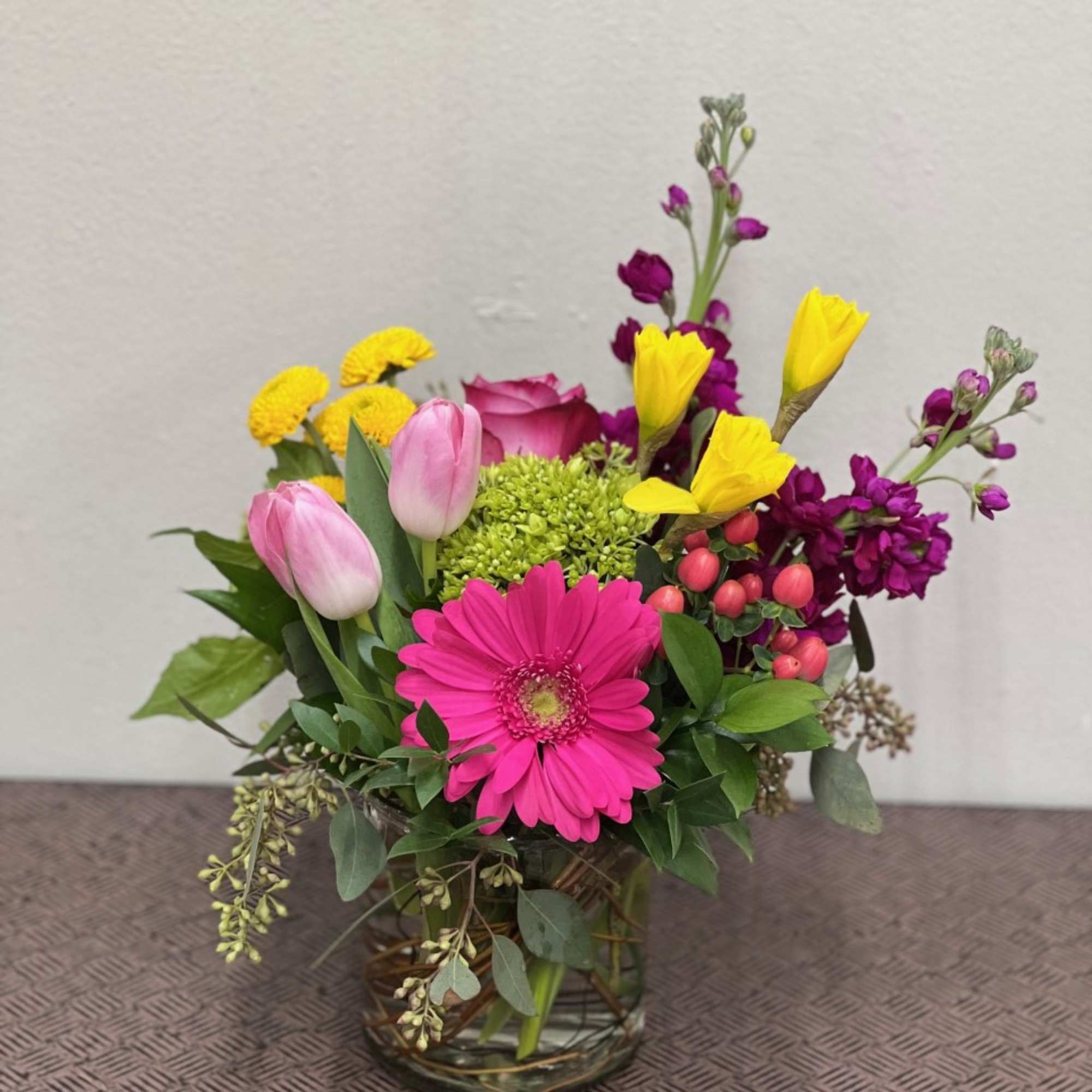 Bright mixed arrangement with pink gerbera, tulips, yellow blooms, and other flowers in a clear glass vase.