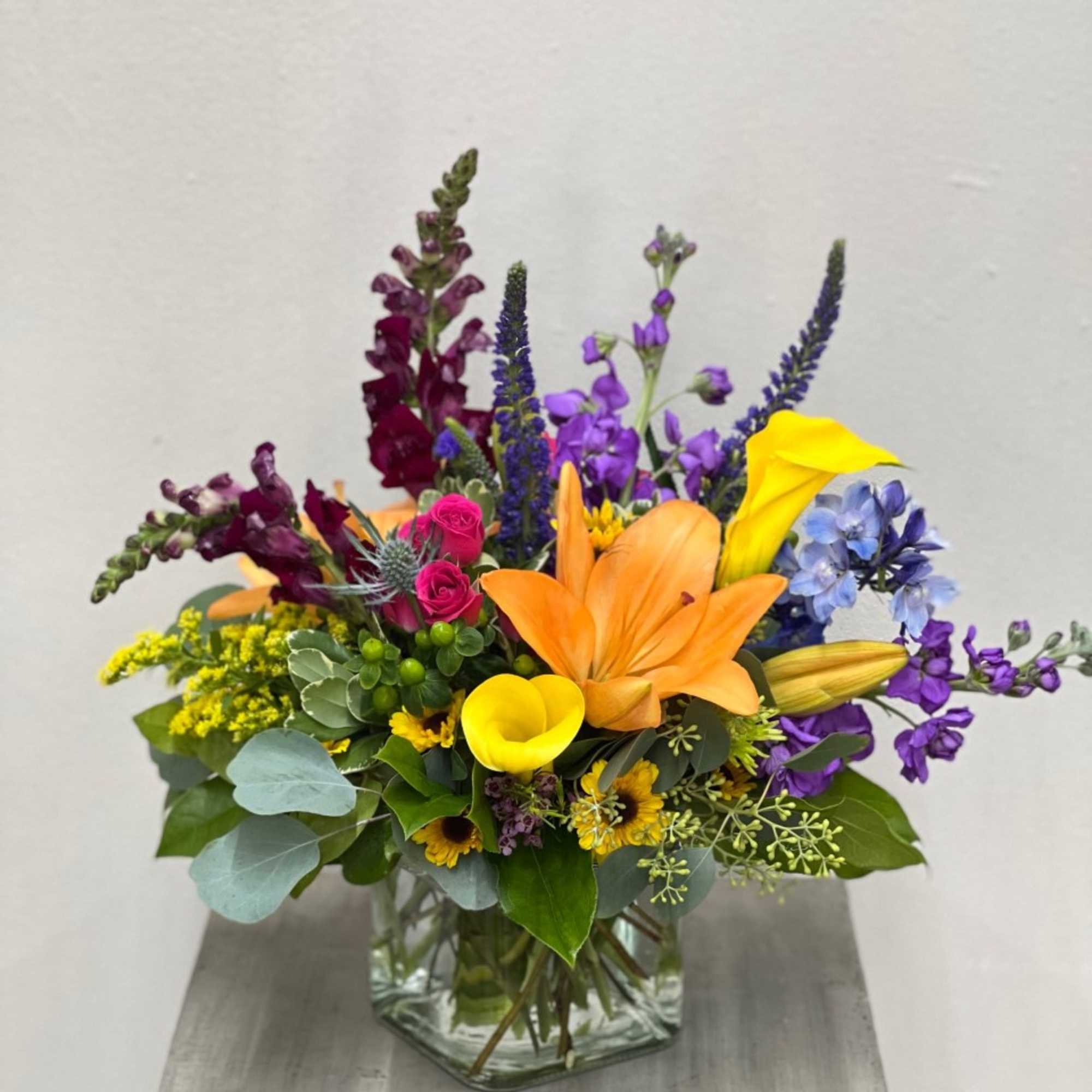 Colorful mixed bouquet with orange lilies, yellow calla lilies, and purple flowers in a clear glass vase