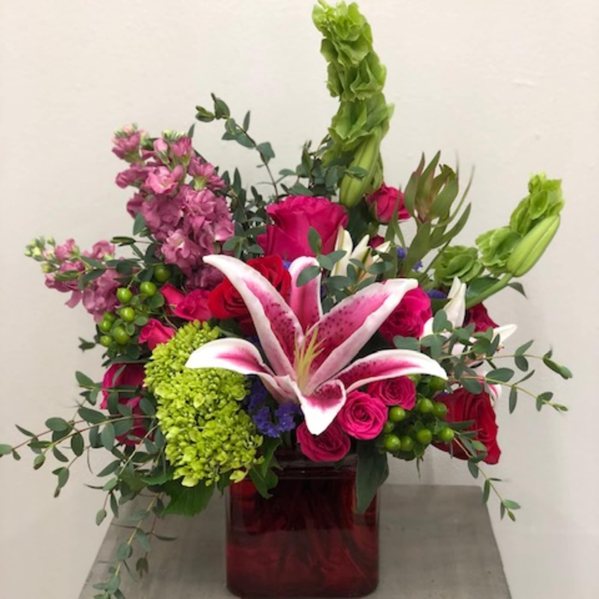 Bright mixed arrangement with pink lilies, roses, green hydrangea and Bells of Ireland in a red glass cube vase