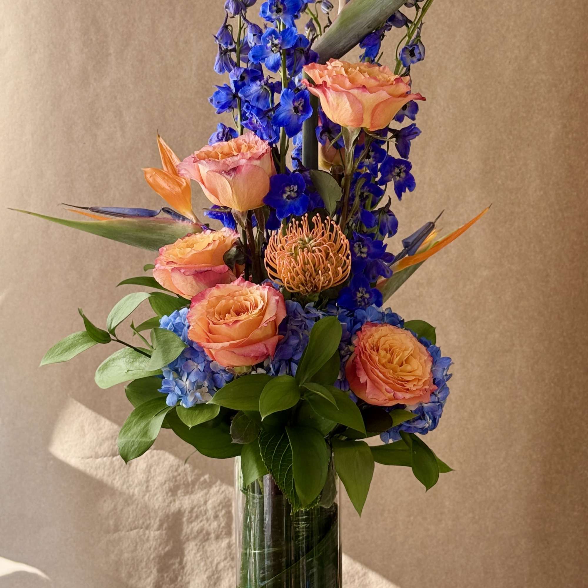 Tall bouquet of orange roses, blue flowers, and bird-of-paradise in a glass vase
