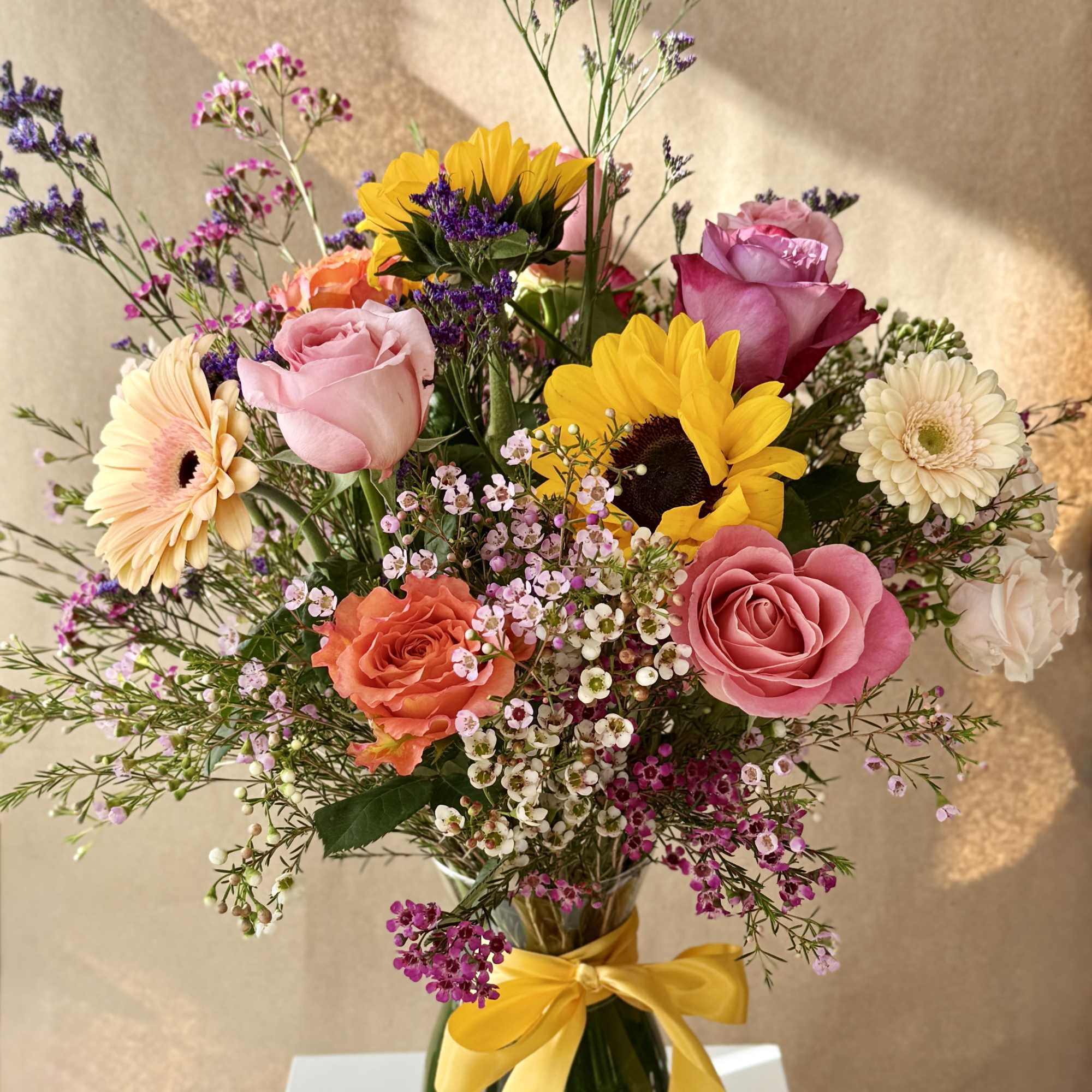 Mixed bouquet of roses, sunflowers, and gerbera daisies in a glass vase with a yellow ribbon