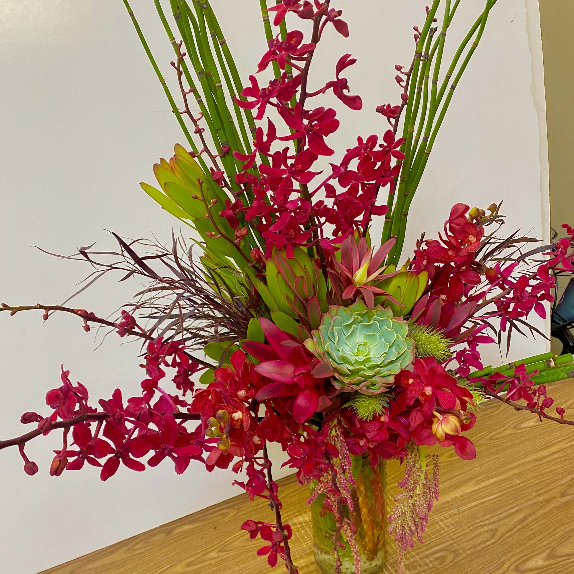 Tall tropical arrangement with red orchids and a succulent in a glass vase