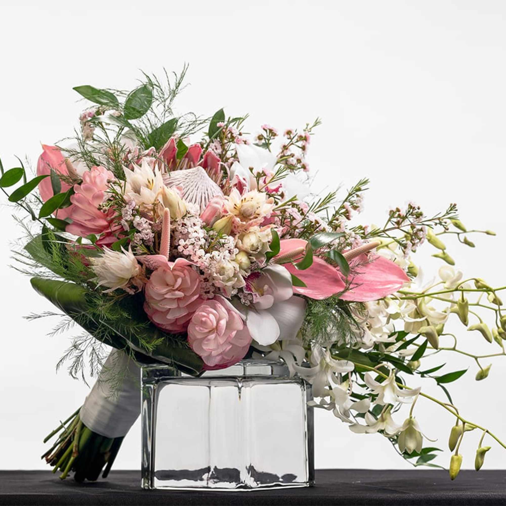 Asymmetrical bouquet of pink tropical flowers, roses, and white orchids resting on a glass vase.